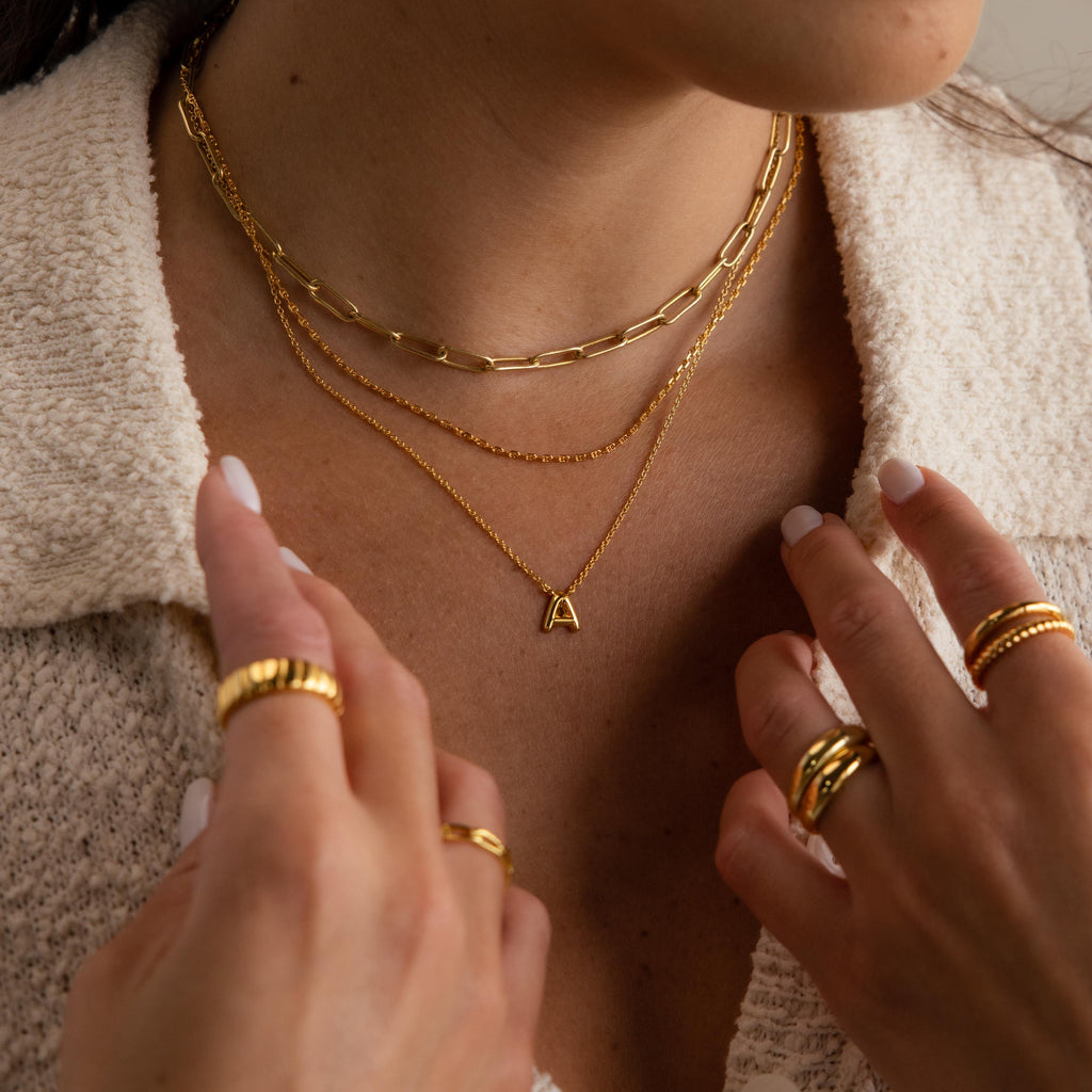 Woman wearing layered gold necklaces, featuring the Dainty Bubble Letter Necklace and a minimalist piece, with gold rings, a textured cream top, and chic white-painted nails.