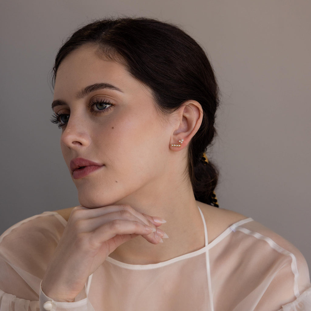 A woman with dark hair, wearing a sheer white top and resting her chin on her hand, showcases the Dainty Pearl Bar Studs.