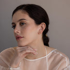 A woman with dark hair, wearing a sheer white top and resting her chin on her hand, showcases the Dainty Pearl Bar Studs.