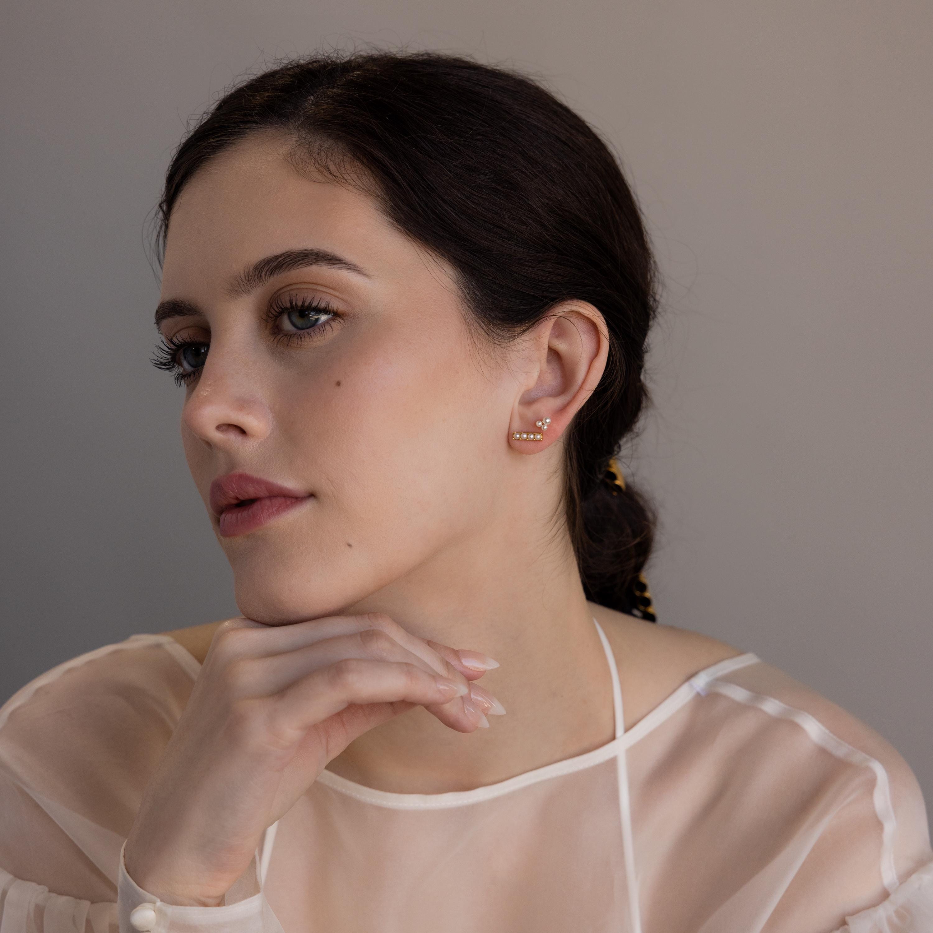 A woman with dark hair, wearing a sheer white top and resting her chin on her hand, showcases the Dainty Pearl Bar Studs.