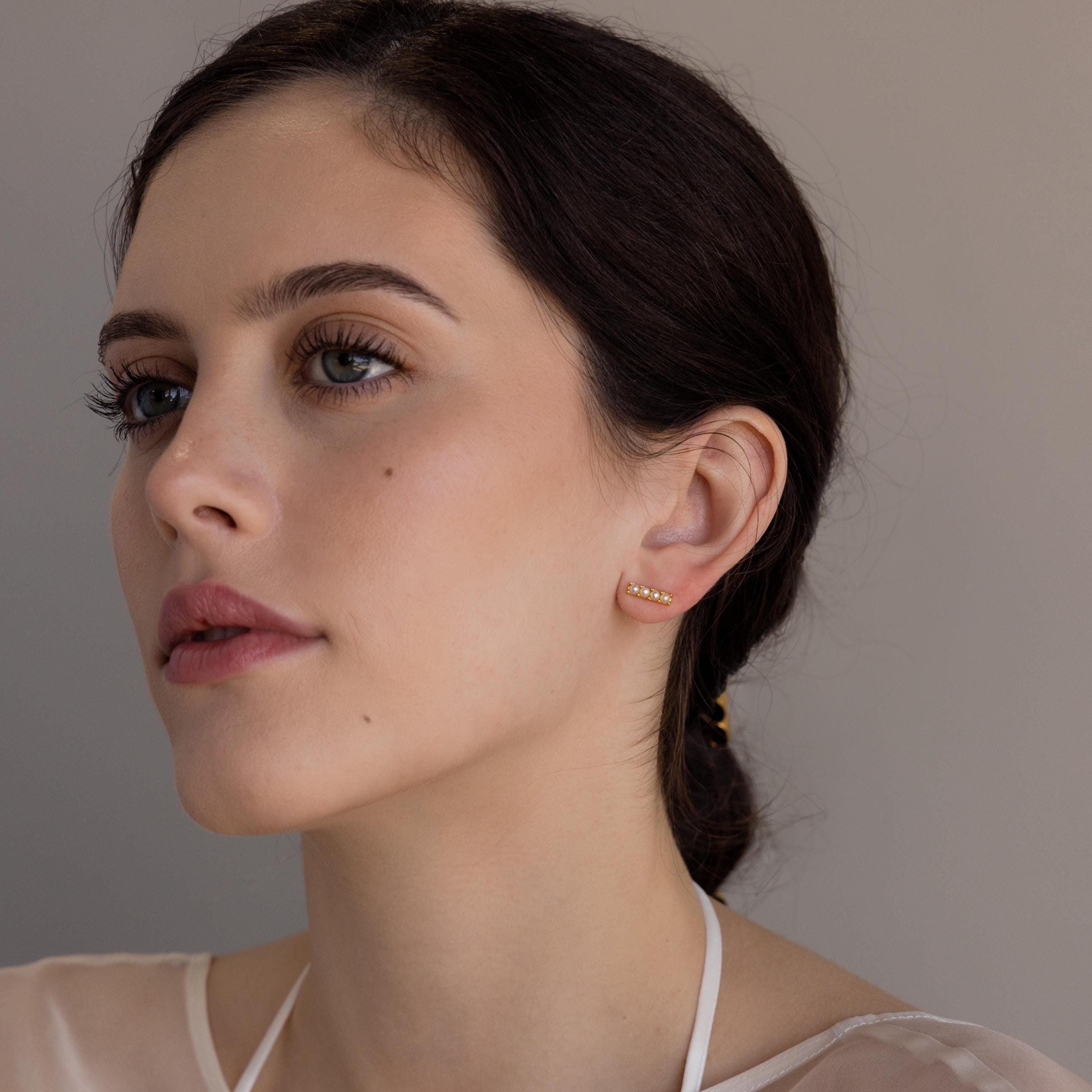 A woman with brown hair wears Dainty Pearl Bar Studs, looking to the side against a neutral background.