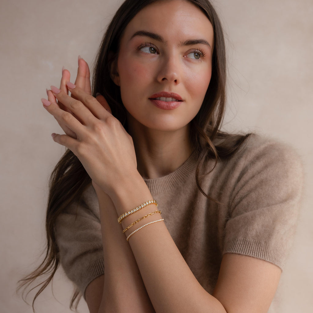 Woman with long brown hair in a beige sweater wears elegant jewelry, featuring the S-Link Diamond Tennis Bracelet in 18K Gold, as she looks to the side.