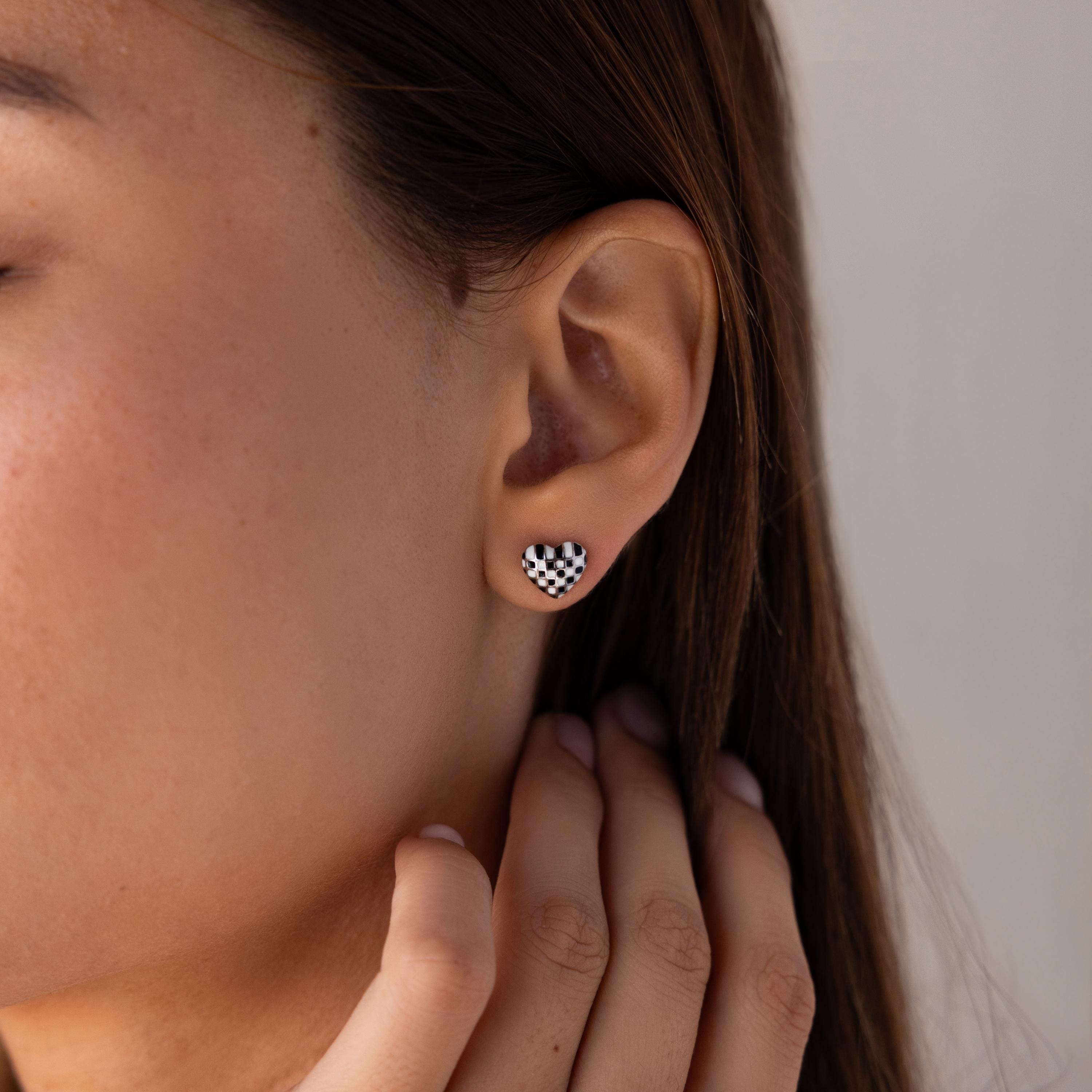 Close-up of a person wearing Checkered Heart Studs, featuring small heart-shaped earrings with a retro black and white checkered pattern.