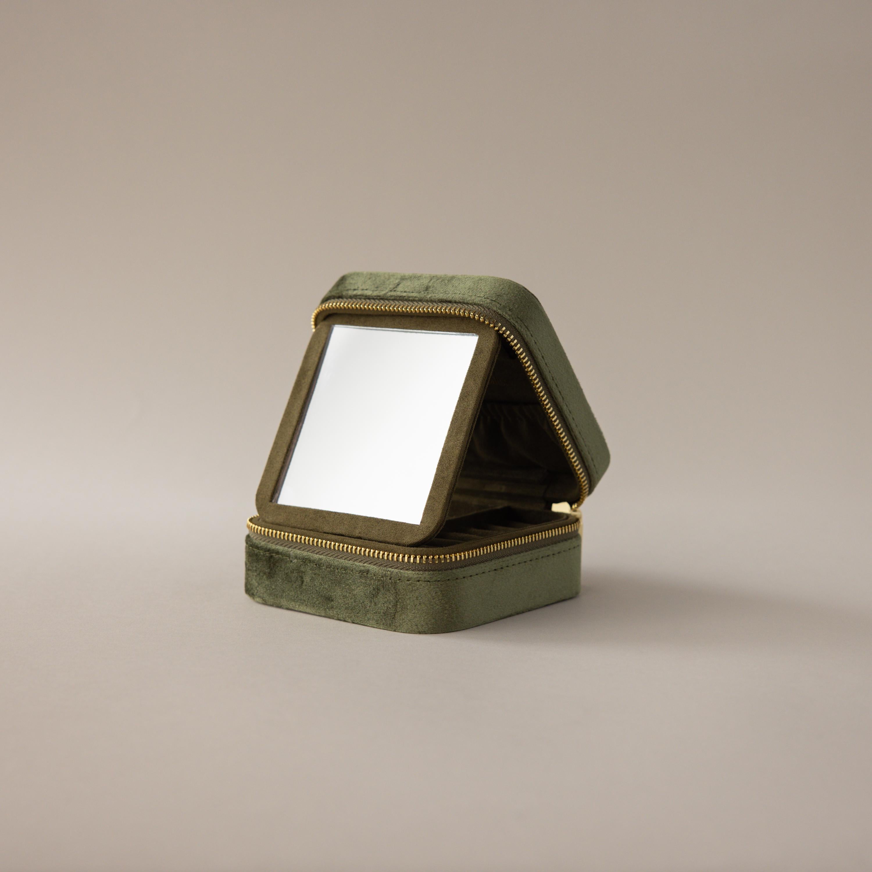 The Velvet Mirror Jewelry Box in green features a square velvet design with a zipper and opens to reveal a mirror—ideal as a compact travel jewelry box, displayed on a plain background.
