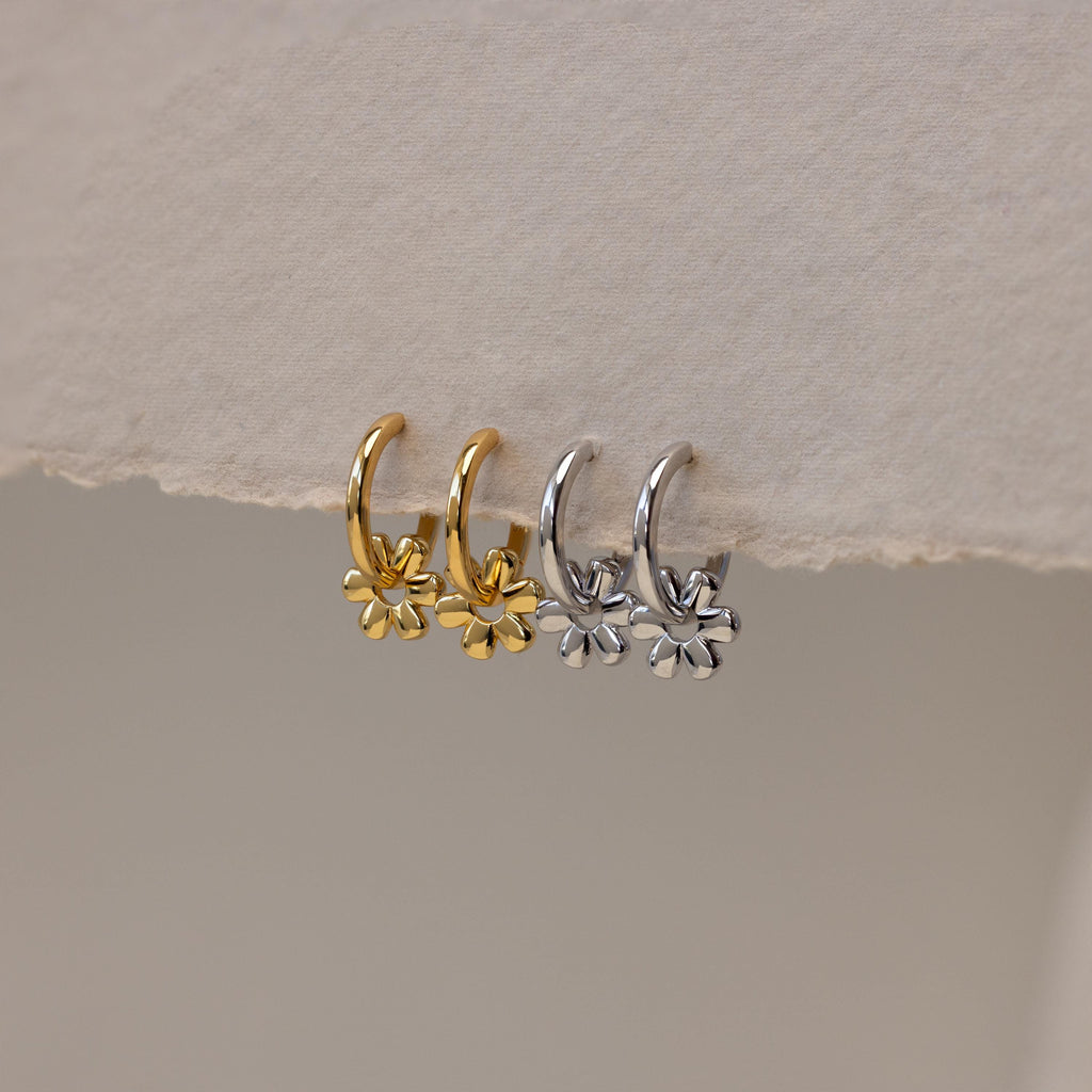 Two pairs of Dangling Blossom Huggies—one gold, one silver—each featuring a delicate flower charm, rest on a textured surface. Perfect for anyone who loves subtle, elegant earrings.