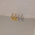 Two pairs of Dangling Blossom Huggies—one gold, one silver—each featuring a delicate flower charm, rest on a textured surface. Perfect for anyone who loves subtle, elegant earrings.