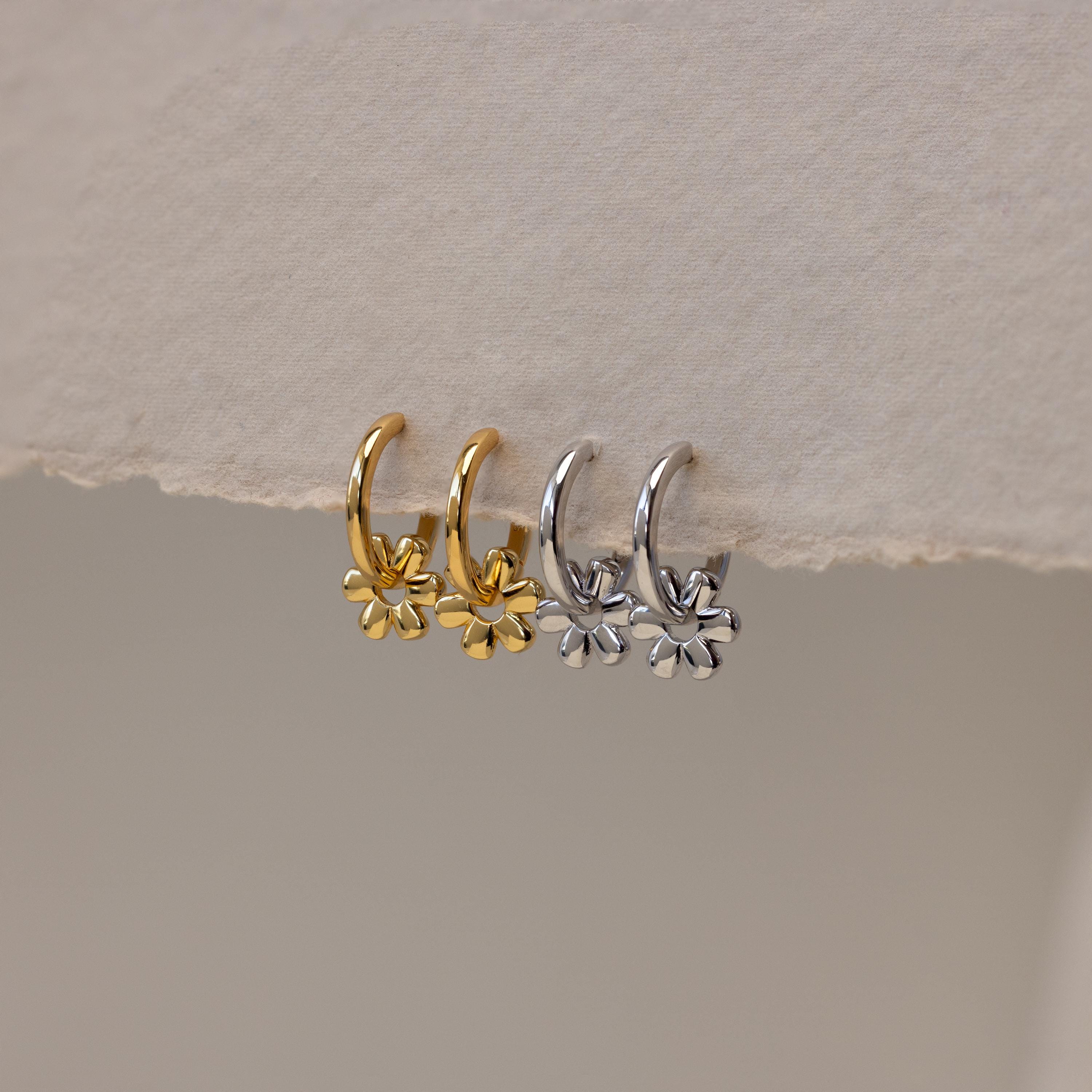 Two pairs of Dangling Blossom Huggies—one gold, one silver—each featuring a delicate flower charm, rest on a textured surface. Perfect for anyone who loves subtle, elegant earrings.