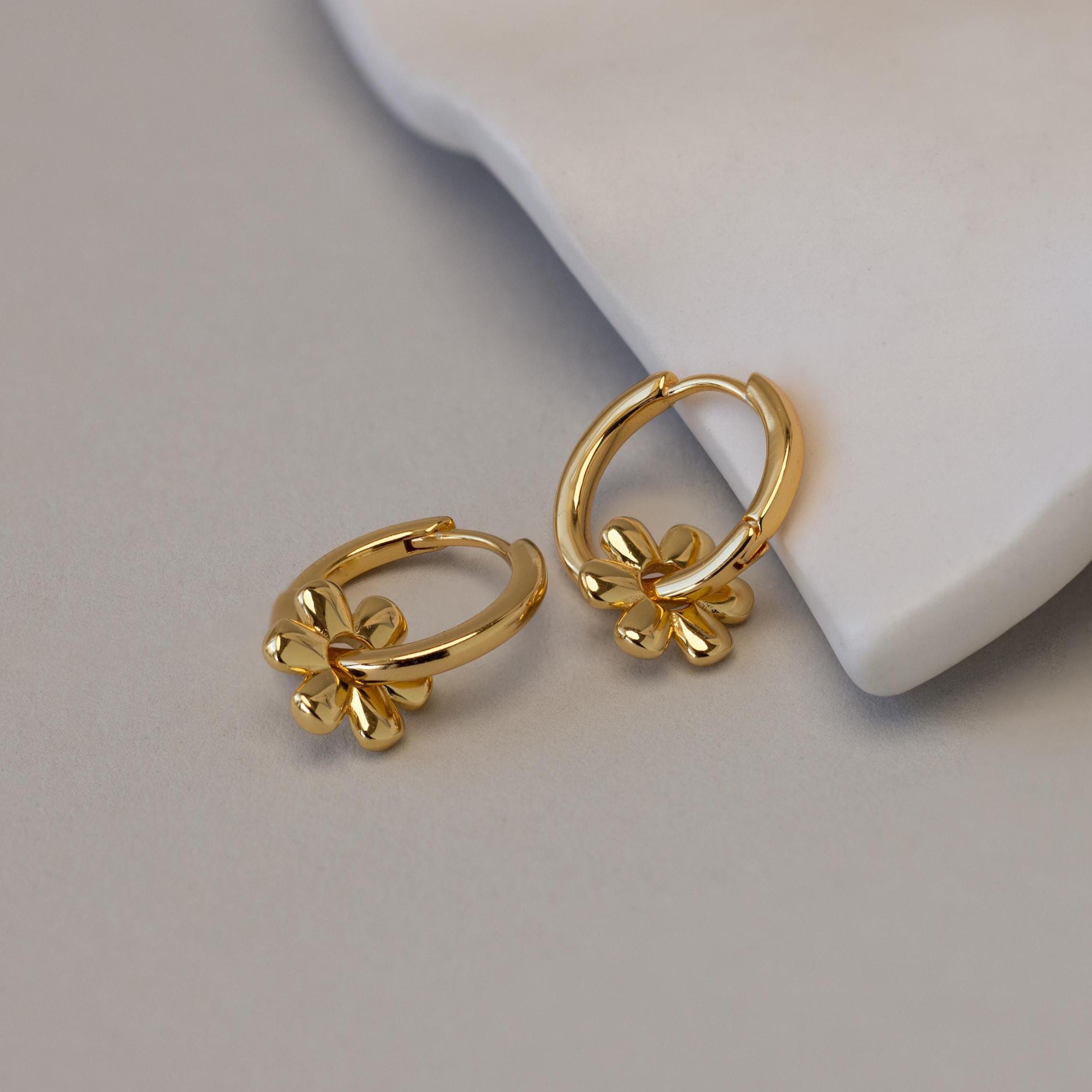 Dangling Blossom Huggies, featuring delicate flower-shaped charms, rest elegantly on a white curved surface and bring a playful twist to timeless gold huggie hoops.
