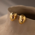 The Bold Disco Earrings, textured gold ovals, are displayed on a ridged beige ceramic vase—perfect for adding retro glam to any look.