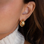 Close-up of a woman's ear wearing the Bold Disco Earrings—shiny, hammered gold hoops with retro glam vibes—paired stylishly with a white knit sweater.