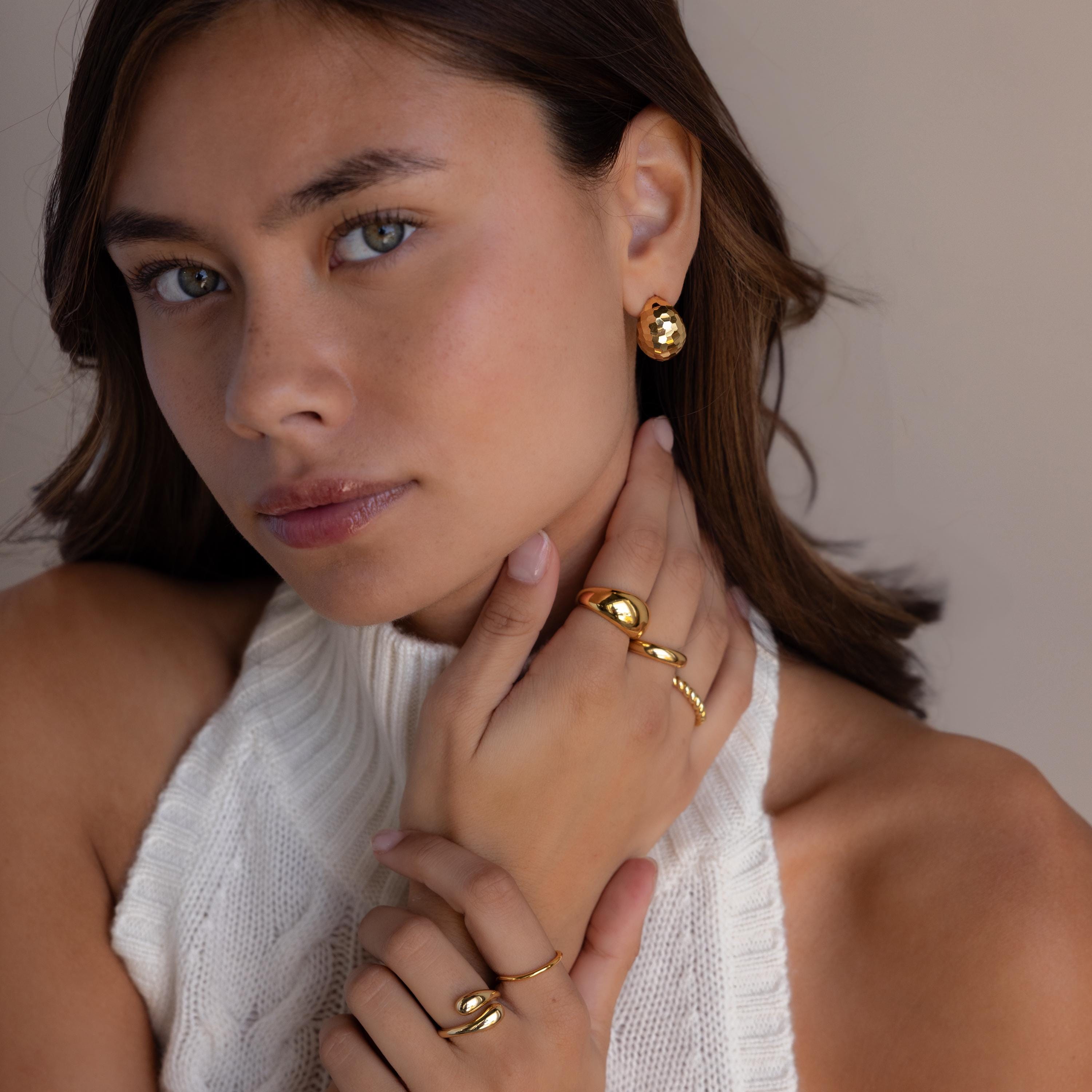 Woman with light eyes in a white knit top wears Bold Disco Earrings—retro glam gold pieces that catch the light as she touches her neck and face.