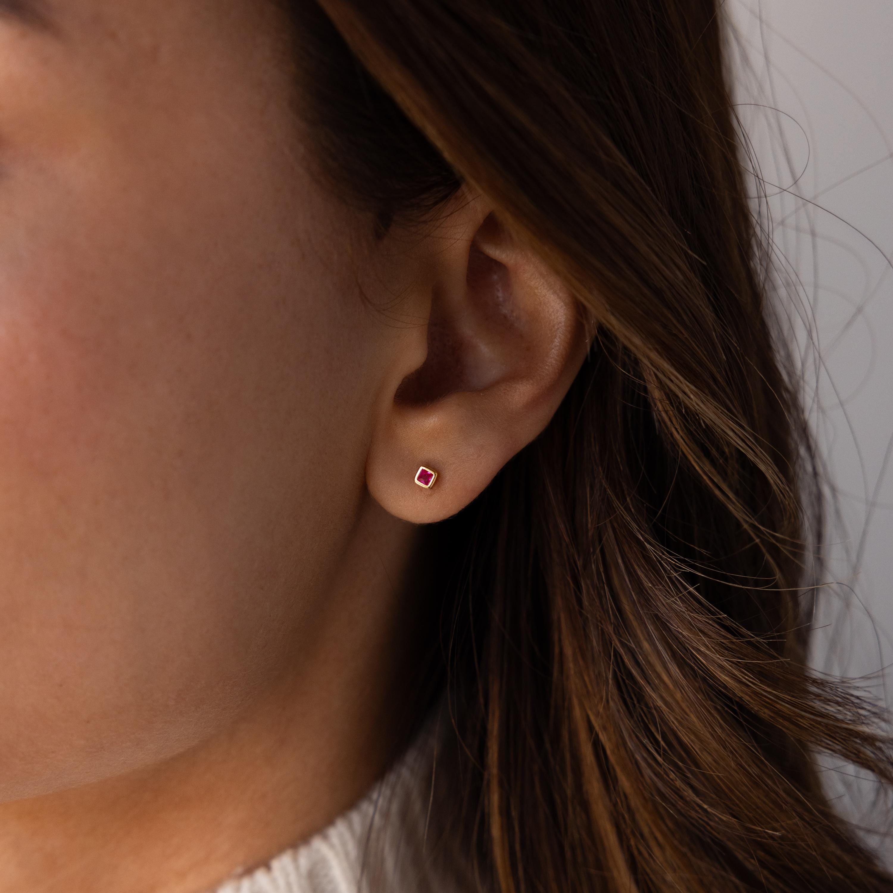 Close-up of a person's ear wearing a Square Bezel Birthstone Flatback Stud; small square red gemstone design and partially visible brown hair.