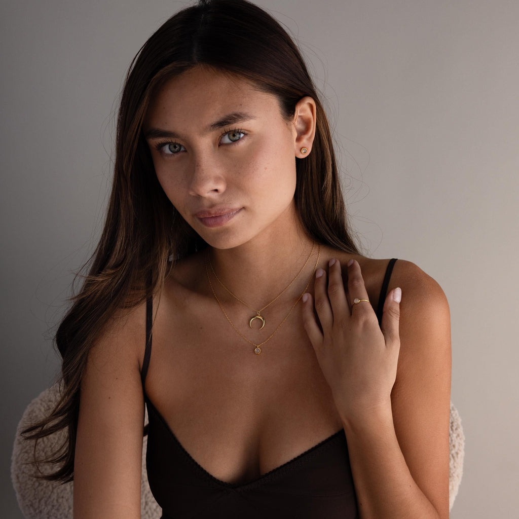 Woman with long brown hair, wearing gold jewelry and a Dainty Round Moonstone Ring, poses in a dark top and looks at the camera with a neutral expression.
