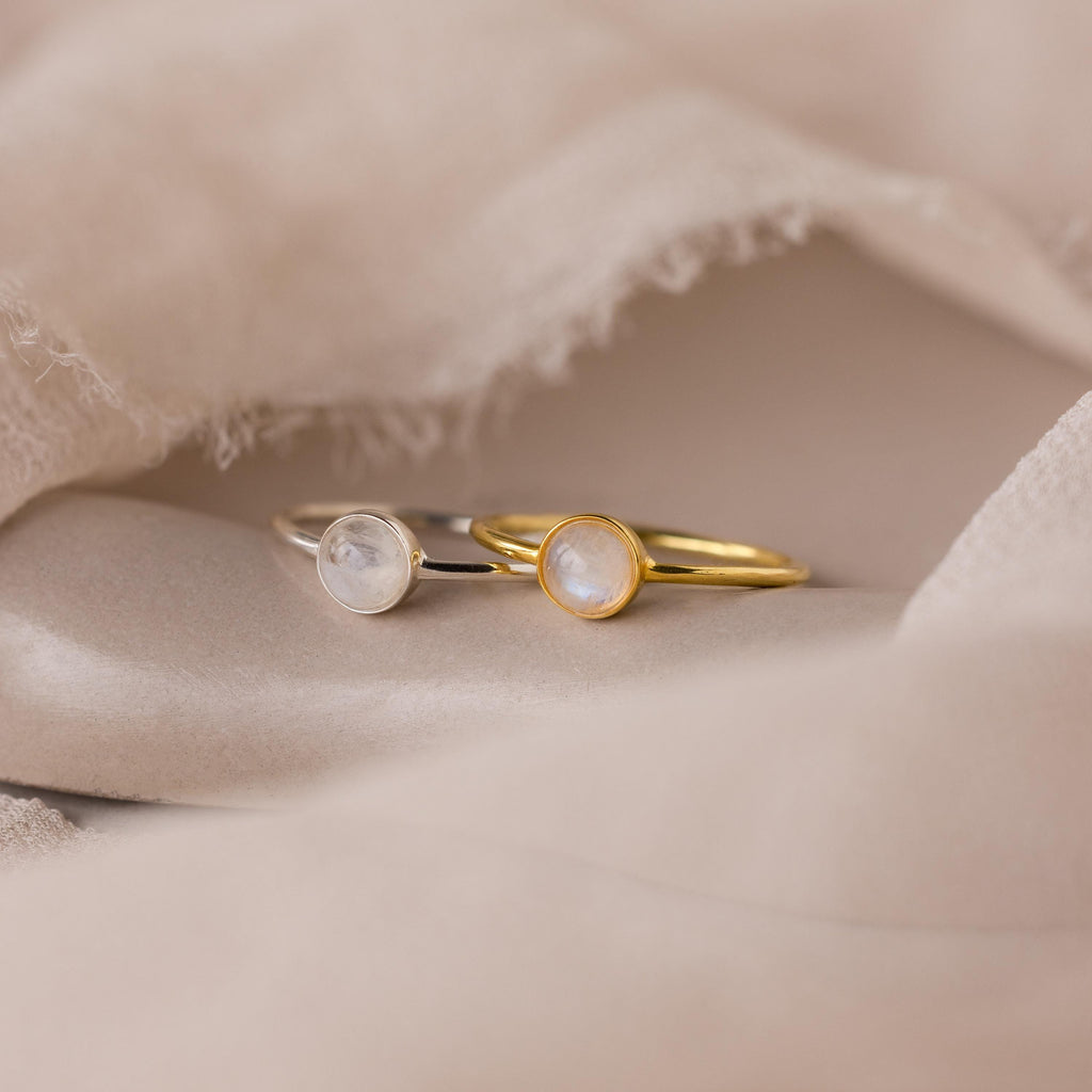 Two delicate Dainty Round Moonstone Rings—one gold, one silver—each featuring a round moonstone, rest on soft beige fabric. They’re the perfect grounding jewelry duo for anyone who loves dainty style.