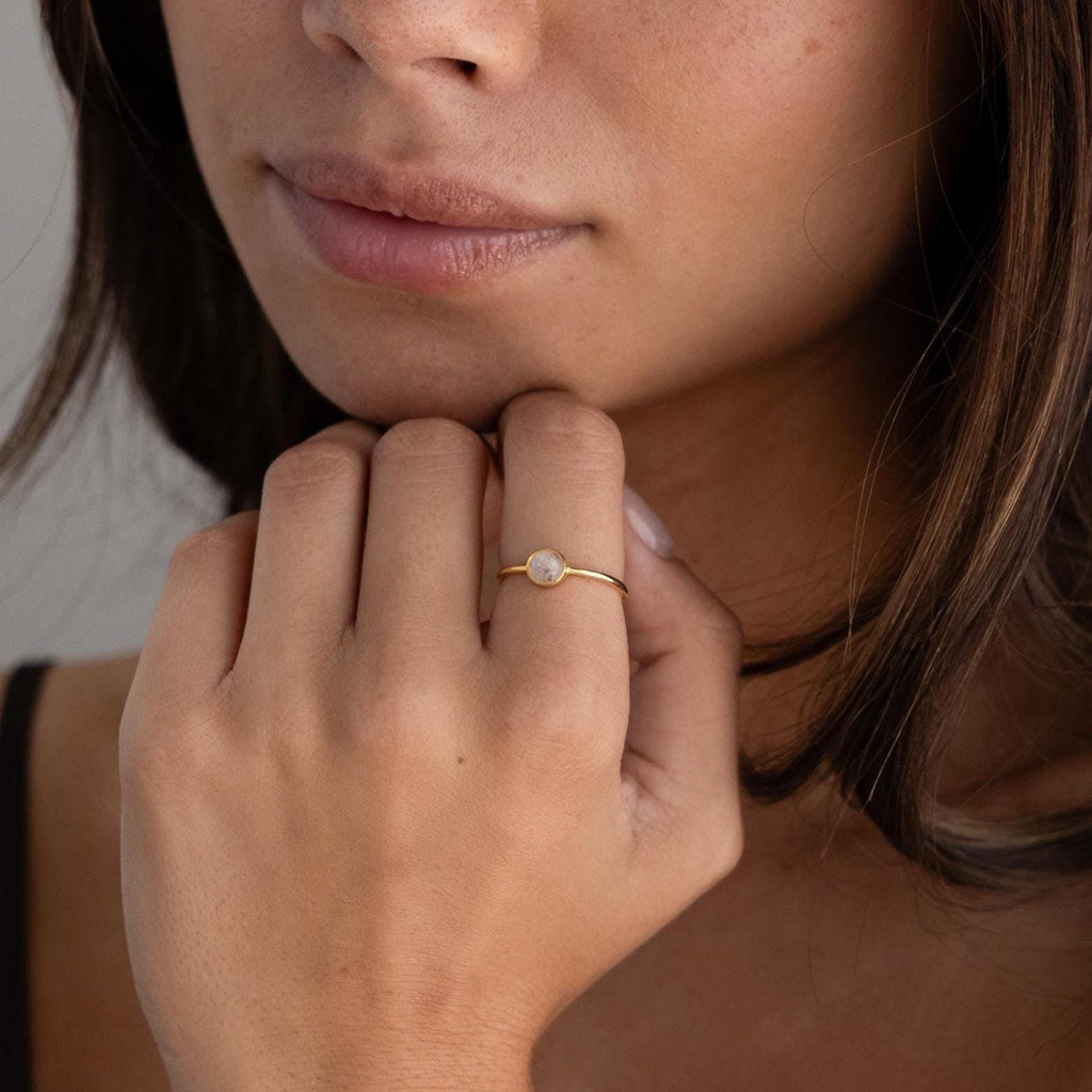 A woman gently touches her chin while wearing the Dainty Round Moonstone Ring, featuring a luminous white stone—an elegant and grounding jewelry piece.