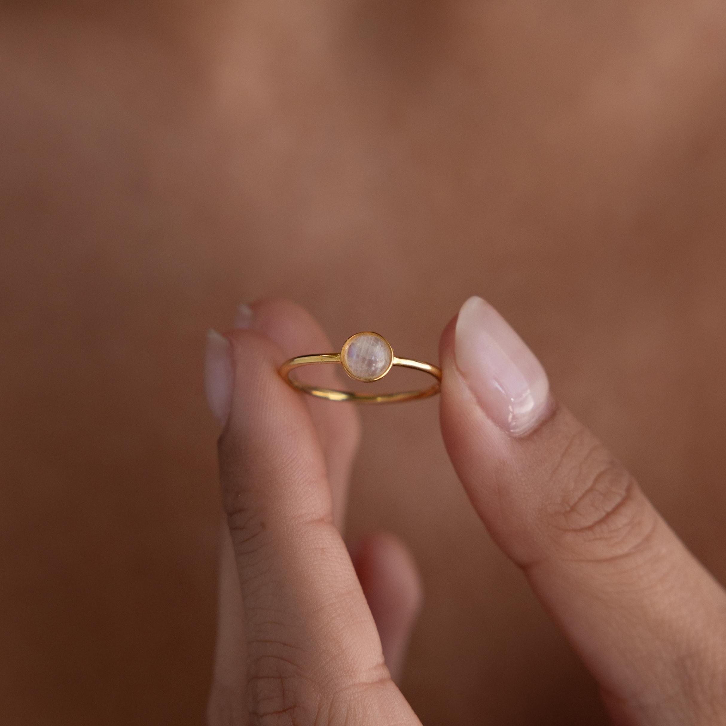 A hand displays the Dainty Round Moonstone Ring, featuring a small, translucent moonstone that catches the light—a perfect piece of grounding jewelry.