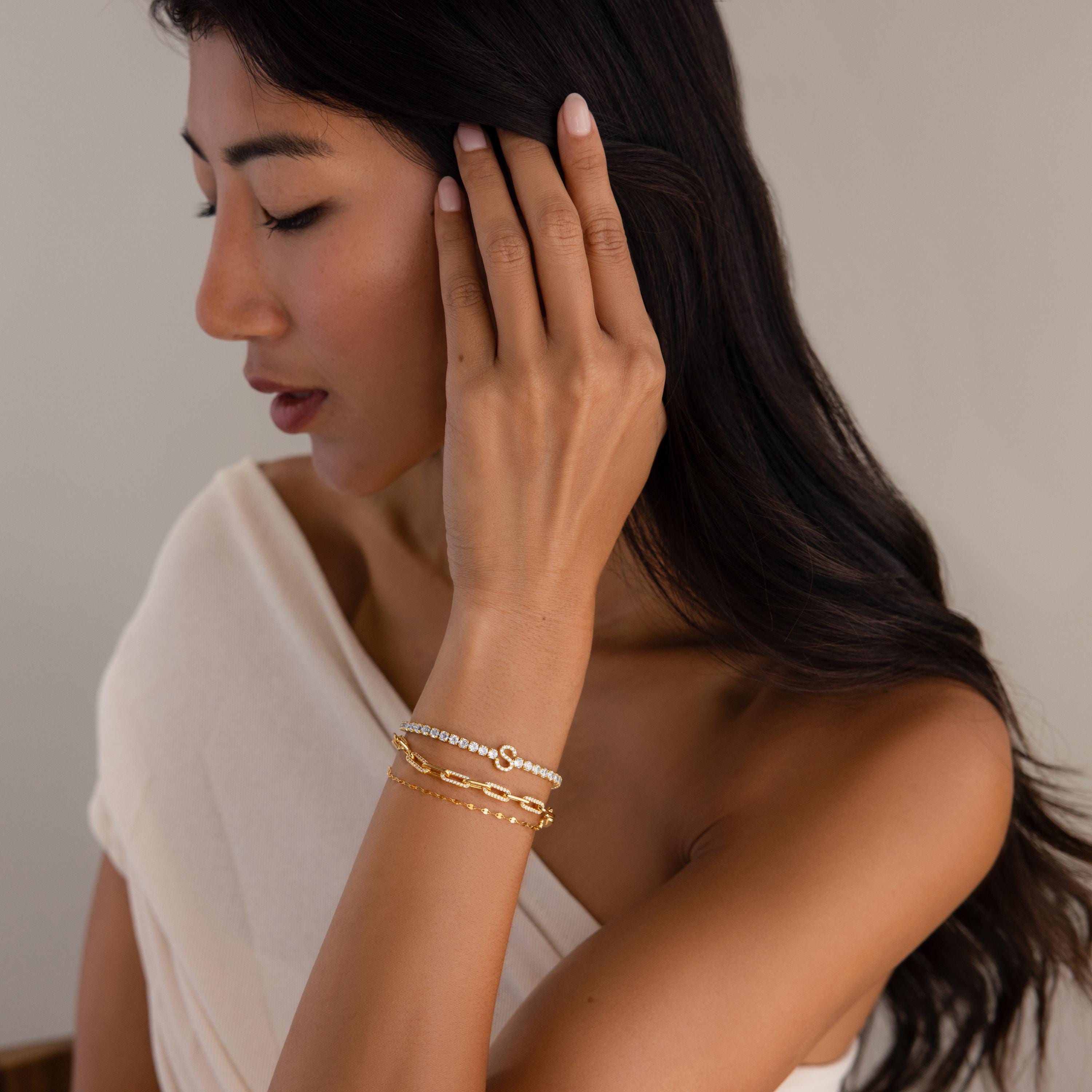 A woman with long dark hair touches her ear, wearing the Diamond Tennis Initial Bracelet stacked with gold and beaded bracelets, paired with an off-shoulder cream top.