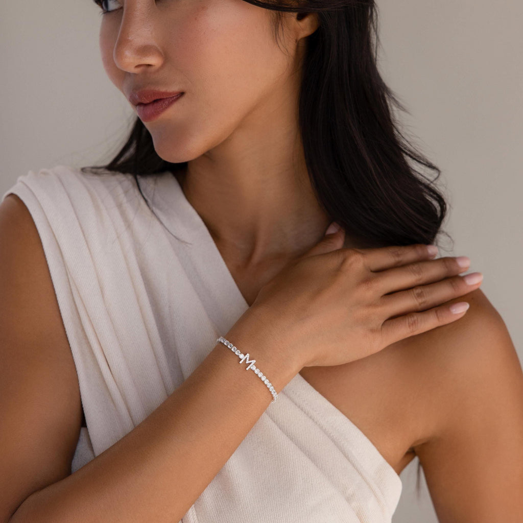 Woman in a white one-shoulder top touches her shoulder while wearing the Diamond Tennis Initial Bracelet.
