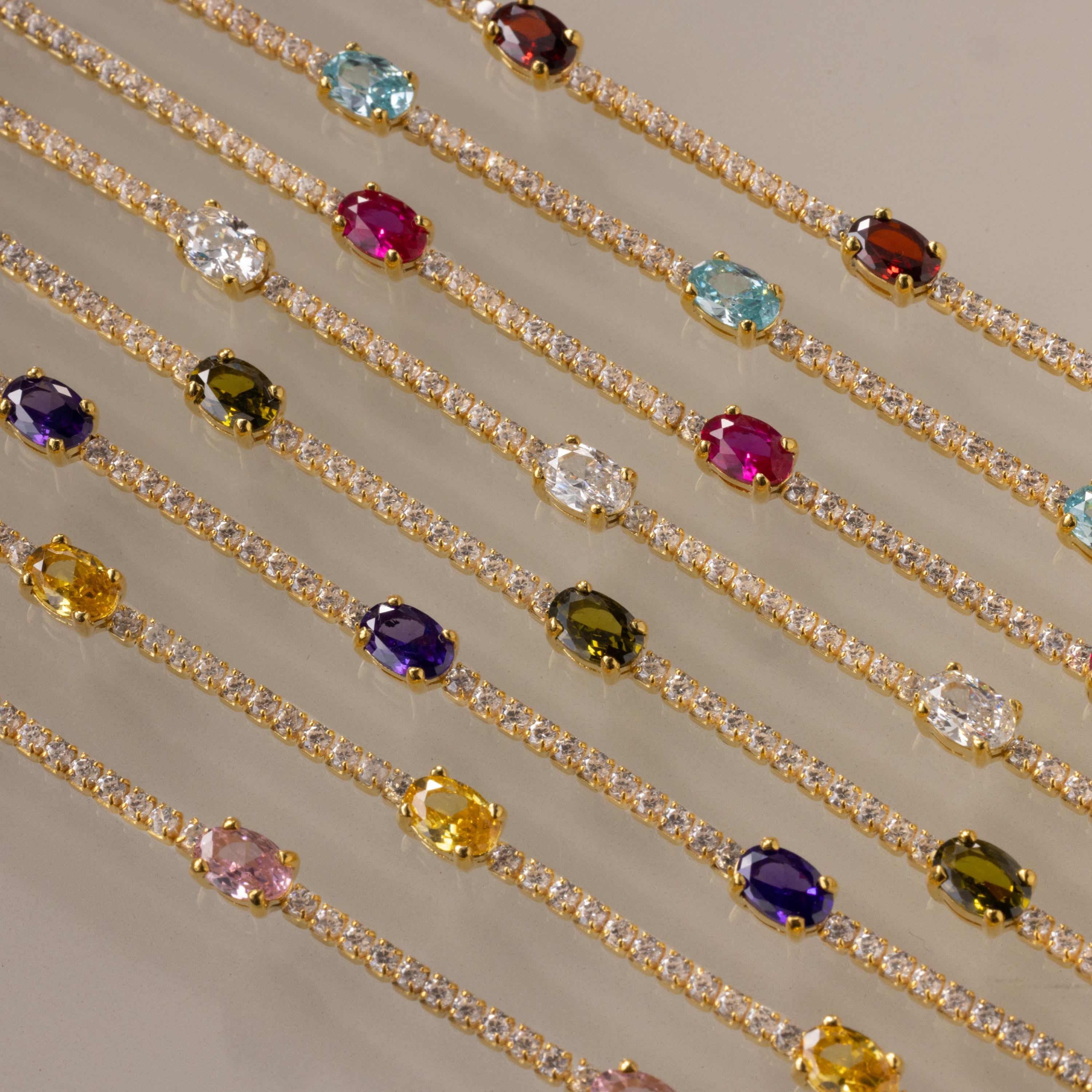 Diamond Birthstone Tennis Bracelets from our collection, adorned with vibrant gemstones and clear crystals, are displayed in neat parallel rows on a beige surface.