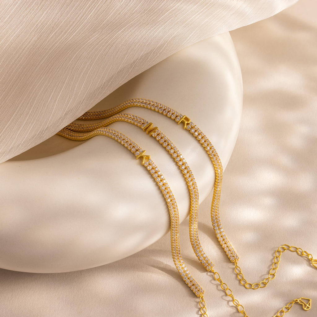 The Initial Duo Tennis Bracelet, a gold and diamond piece with geometric charms and a custom initial design, is elegantly displayed on a smooth, cream-colored surface.