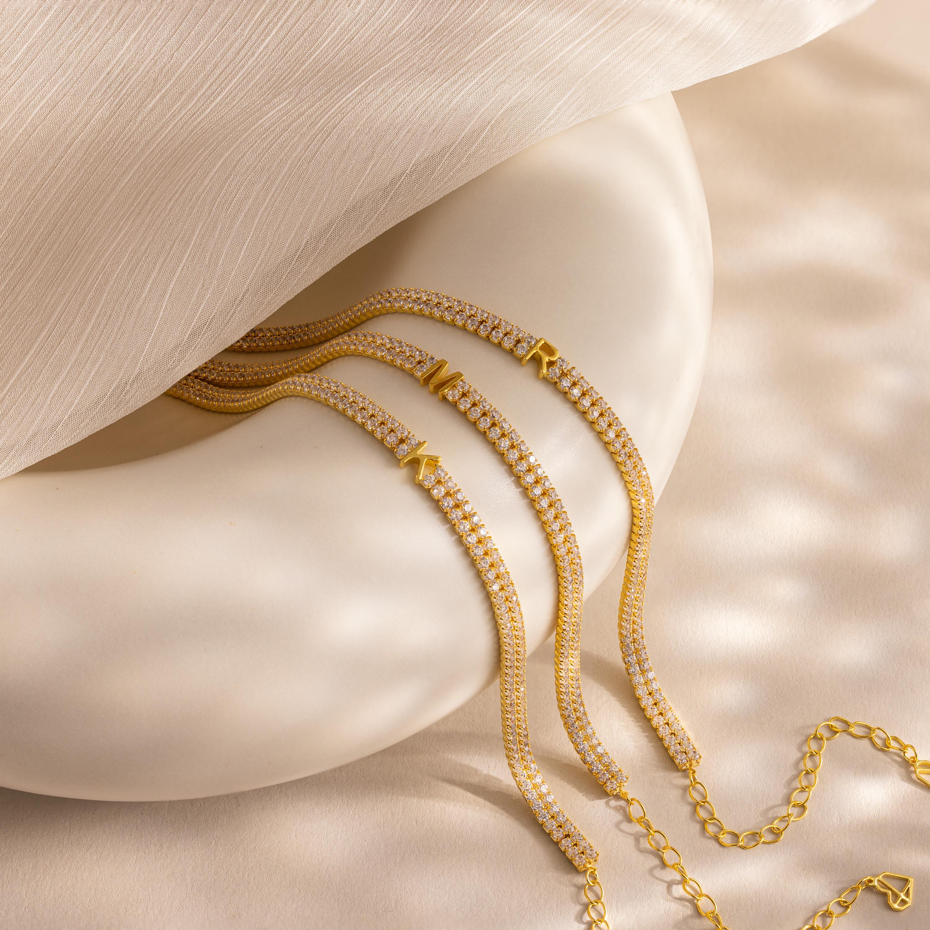 The Initial Duo Tennis Bracelet, a gold and diamond piece with geometric charms and a custom initial design, is elegantly displayed on a smooth, cream-colored surface.