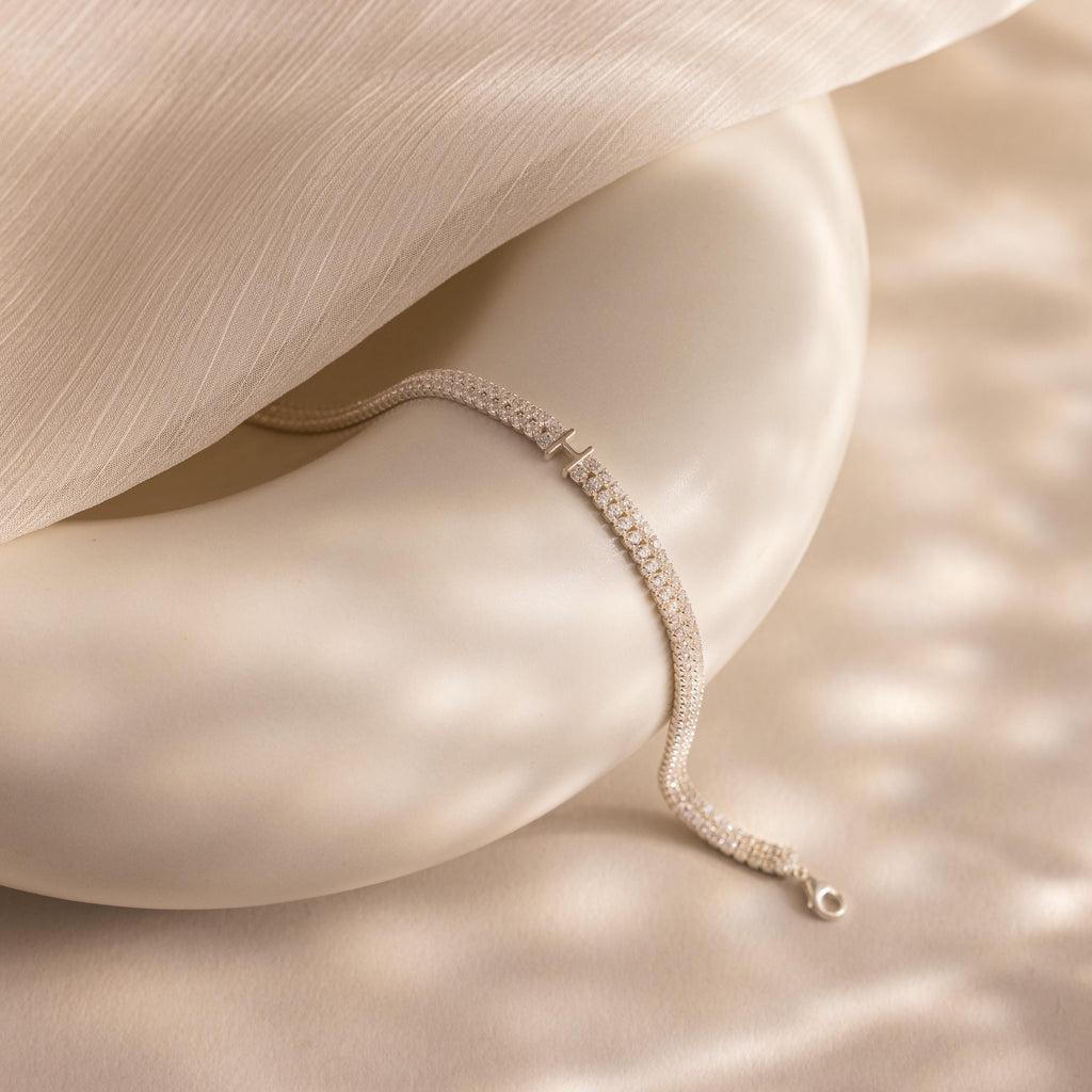 The Initial Duo Tennis Bracelet is elegantly displayed on a smooth white sculptural object, accented by soft beige fabric overhead.