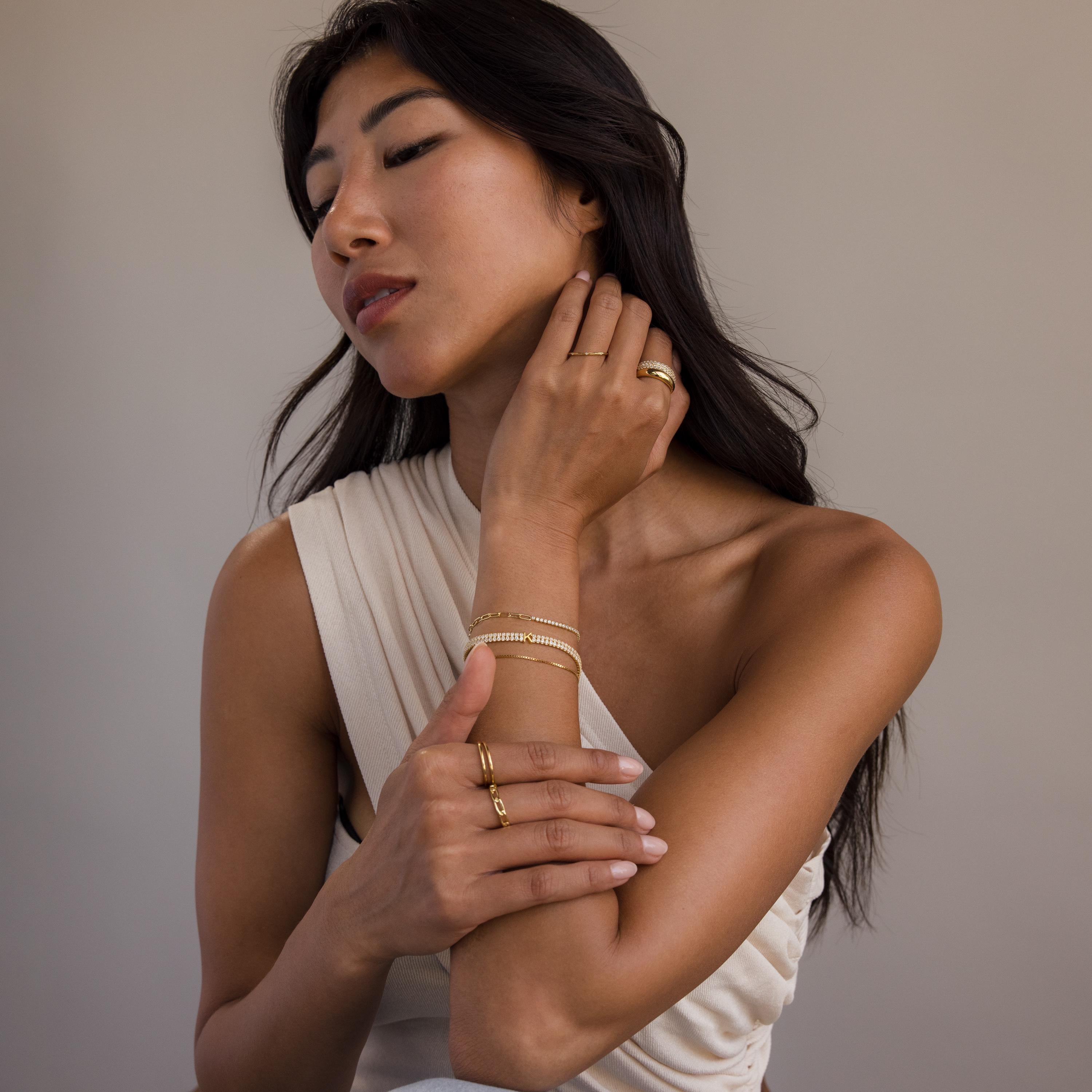 A woman in a sleeveless white top wears gold rings and the Initial Duo Tennis Bracelet, posing with her hand on her neck and arm.
