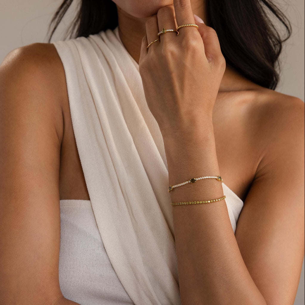 Woman in a white sleeveless top wears gold rings and our Diamond Birthstone Tennis Bracelet, hand near her face.