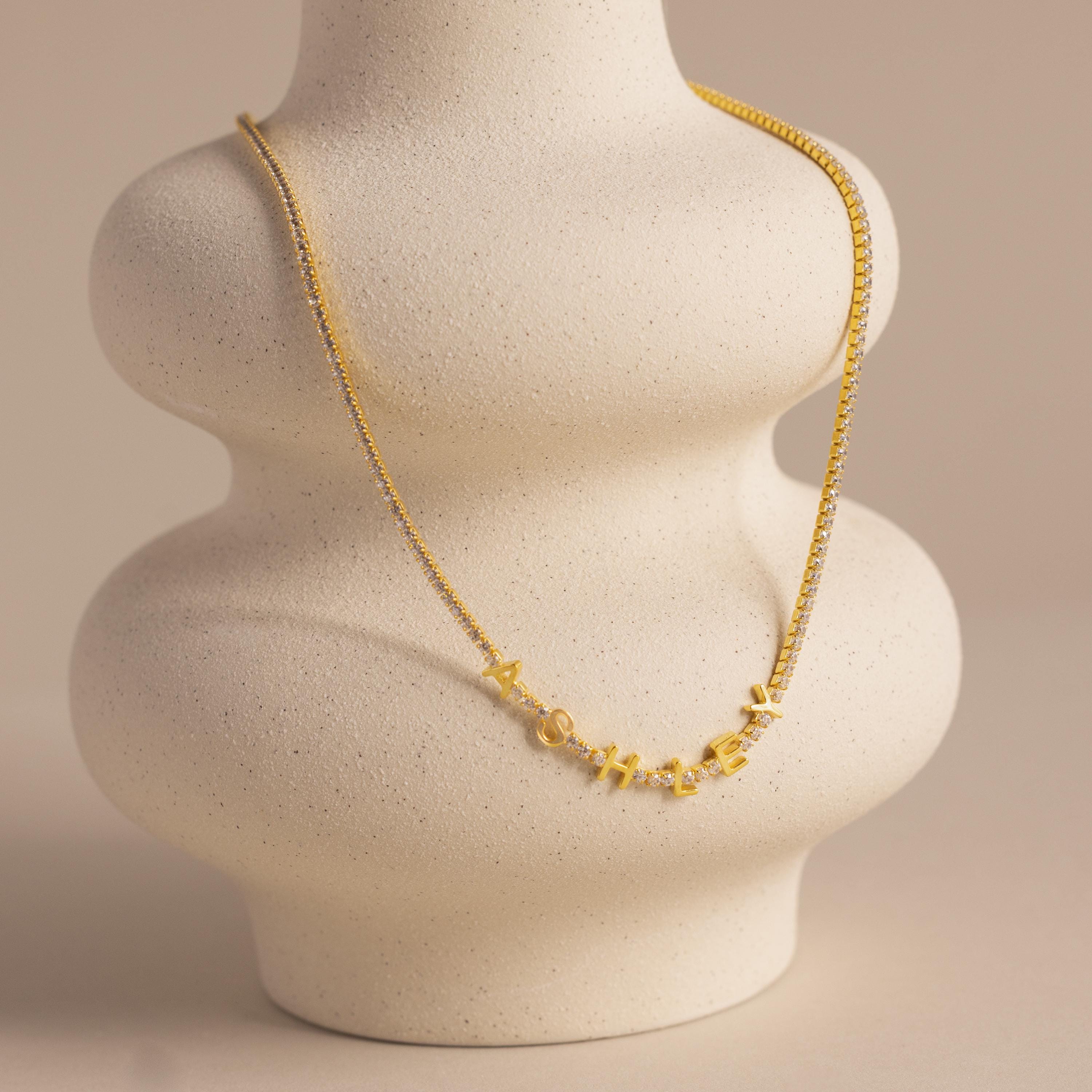The Diamond Tennis Letter Necklace, spelling “Mother,” is elegantly displayed on a curvy, light beige ceramic bust—an ideal, sparkling tribute to celebrate mom in timeless style.