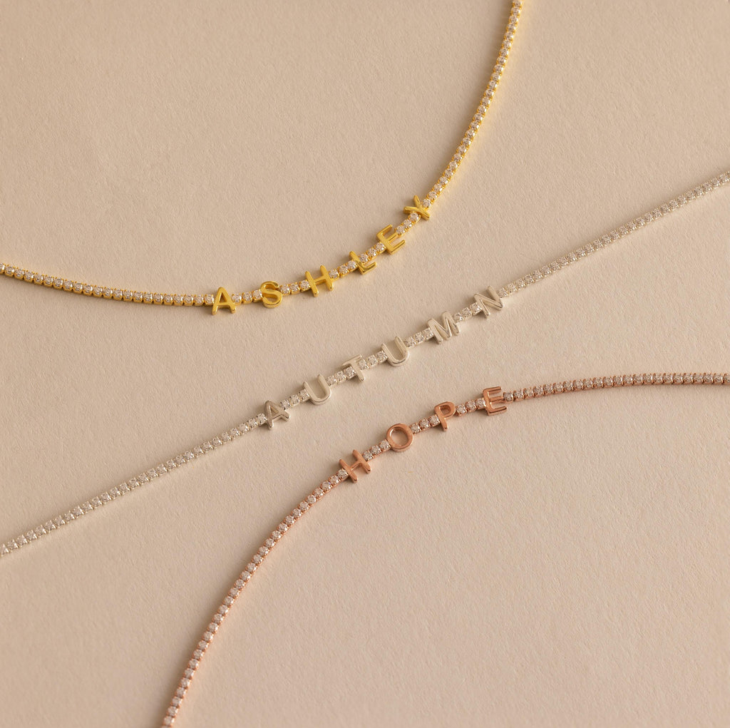 Three Diamond Tennis Letter Necklaces spelling ASHLEY, AUTUMN, and HOPE in gold, silver, and rose gold are showcased on a beige background.