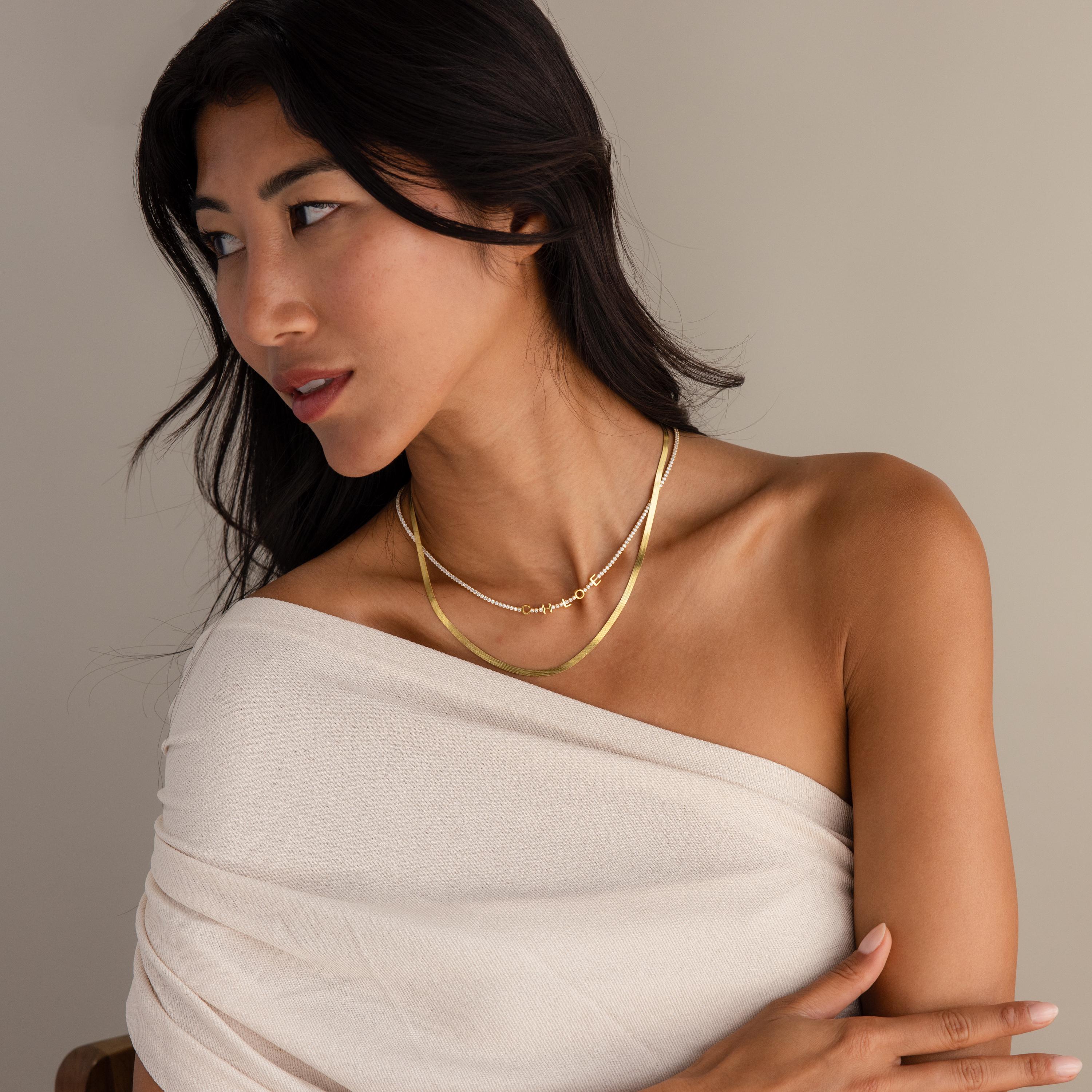 Woman in an off-shoulder white top wearing layered gold necklaces, including the Diamond Tennis Letter Necklace, and looking to the side.