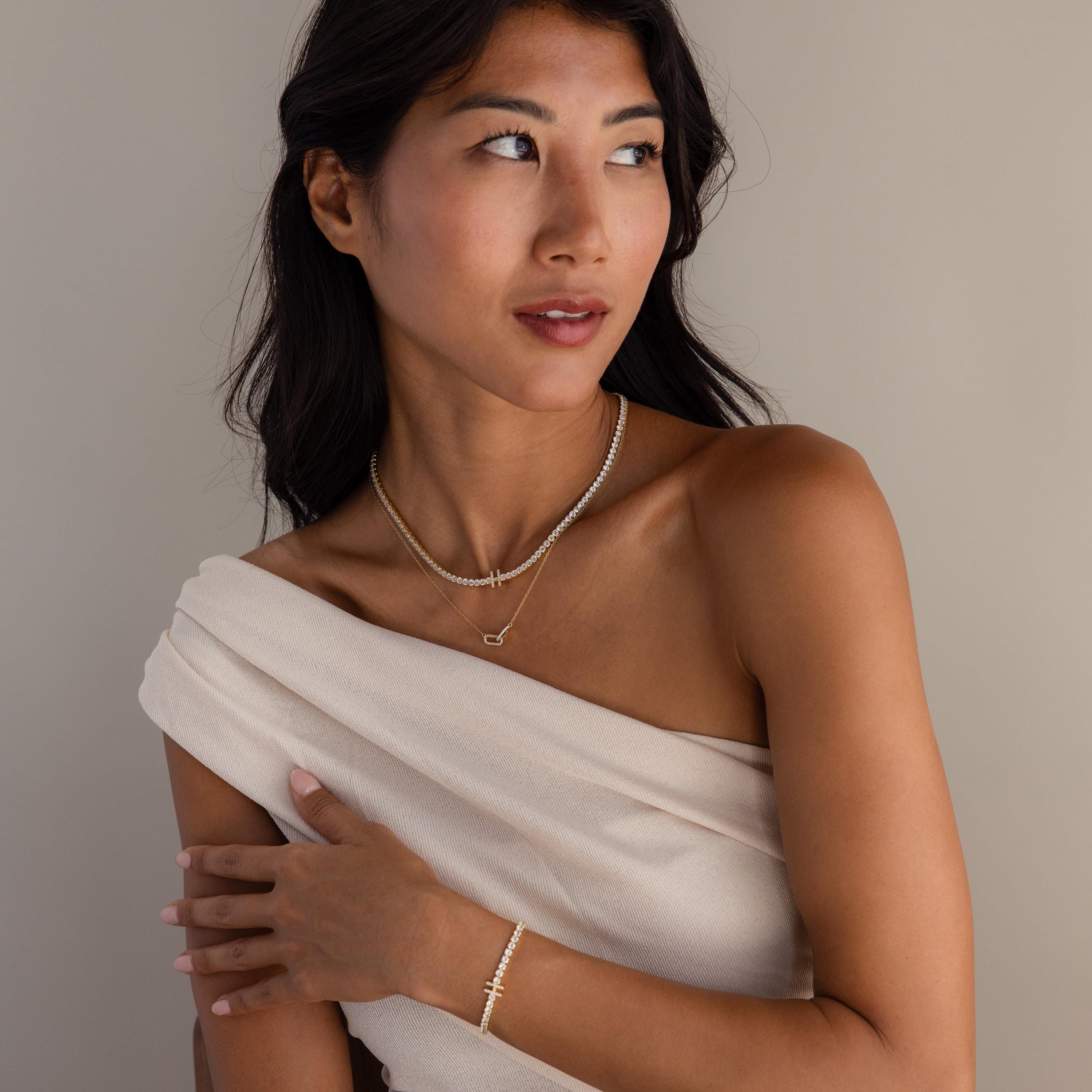Woman in an off-shoulder beige top wearing a gold bracelet and layered gold necklaces, including the Diamond Tennis Initial Necklace, looking to the side.
