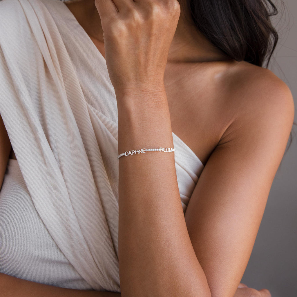 A woman wears a white top and the Multiple Name Tennis Bracelet, featuring custom name charms "DAPHNE" and "PALOMA" on her wrist.