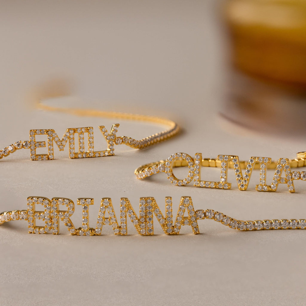 Three gold Pave Name Tennis Bracelets, each personalized with diamond-studded names—EMILY, OLIVIA, and BRIANNA—are displayed on a beige surface, showcasing the elegant and timeless charm of the Pave Name Tennis Bracelet.