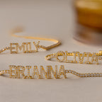 Three gold Pave Name Tennis Bracelets, each personalized with diamond-studded names—EMILY, OLIVIA, and BRIANNA—are displayed on a beige surface, showcasing the elegant and timeless charm of the Pave Name Tennis Bracelet.