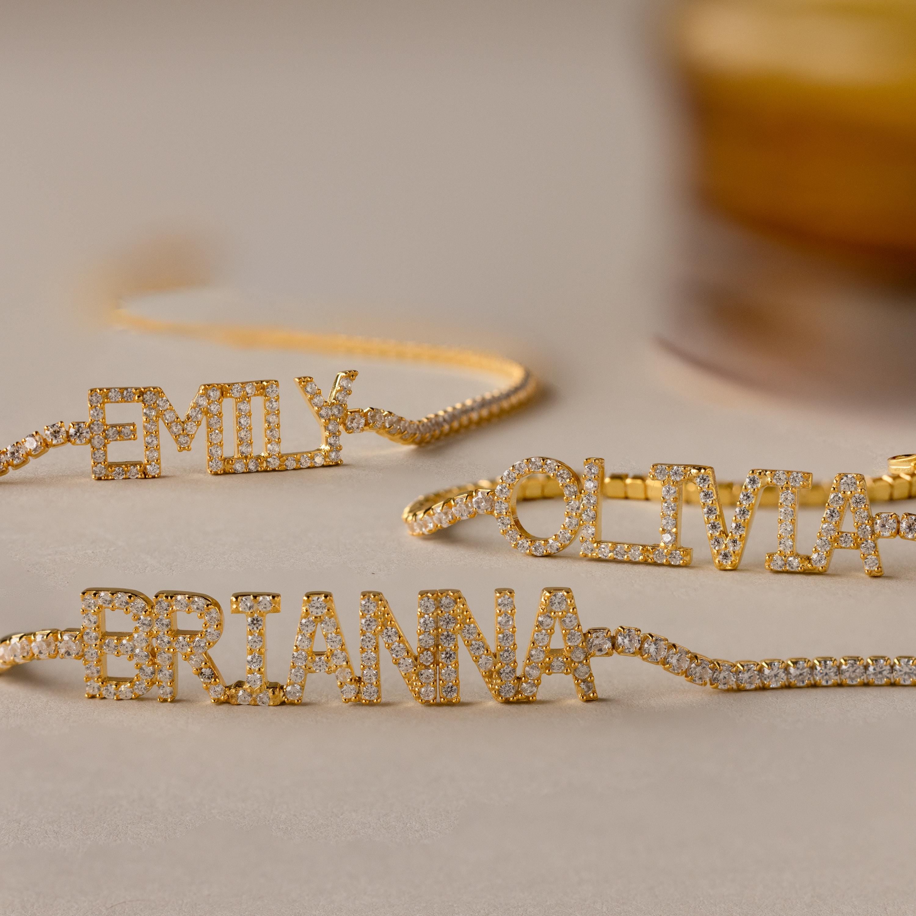 Three gold Pave Name Tennis Bracelets, each personalized with diamond-studded names—EMILY, OLIVIA, and BRIANNA—are displayed on a beige surface, showcasing the elegant and timeless charm of the Pave Name Tennis Bracelet.