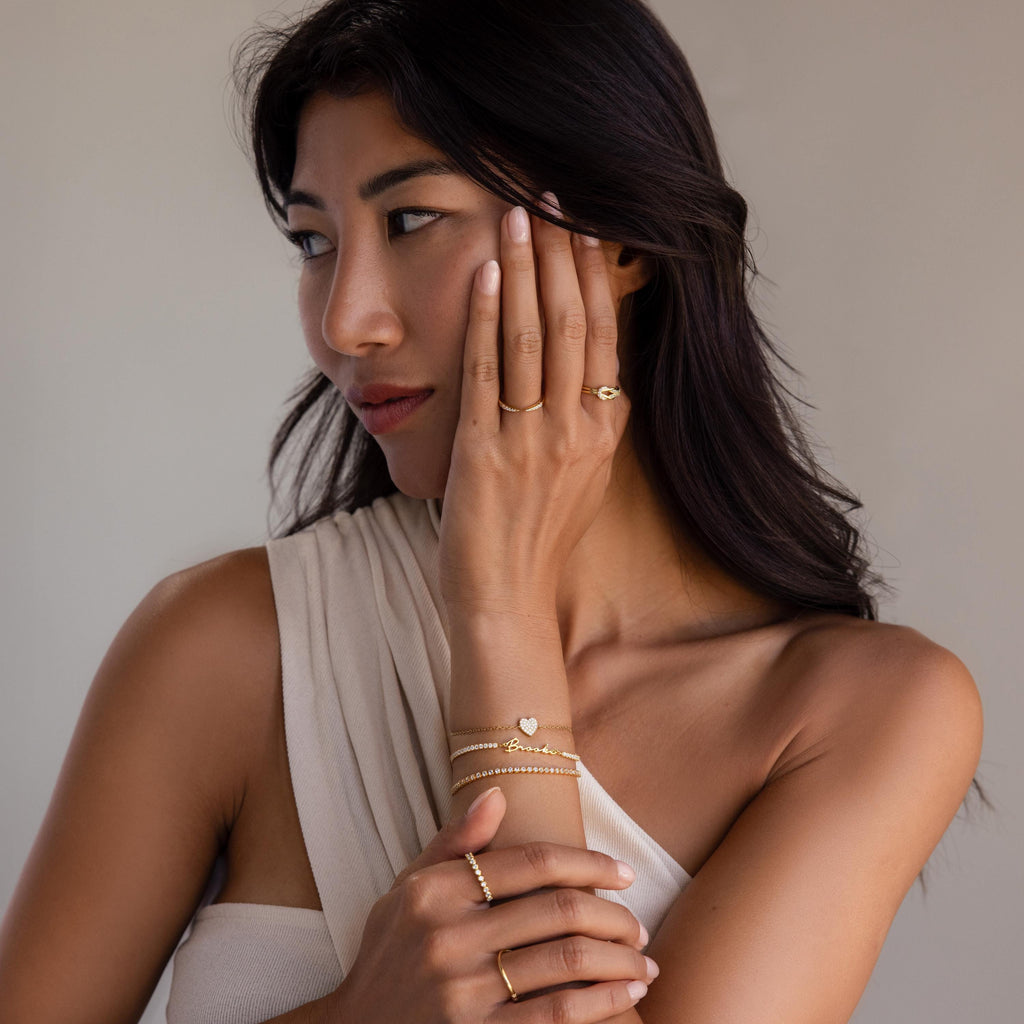 A woman wearing a white one-shoulder top and gold rings showcases the Venice Name Tennis Bracelet as she gazes to the side, her hand gently resting on her face.