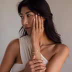 A woman wearing a white one-shoulder top and gold rings showcases the Venice Name Tennis Bracelet as she gazes to the side, her hand gently resting on her face.