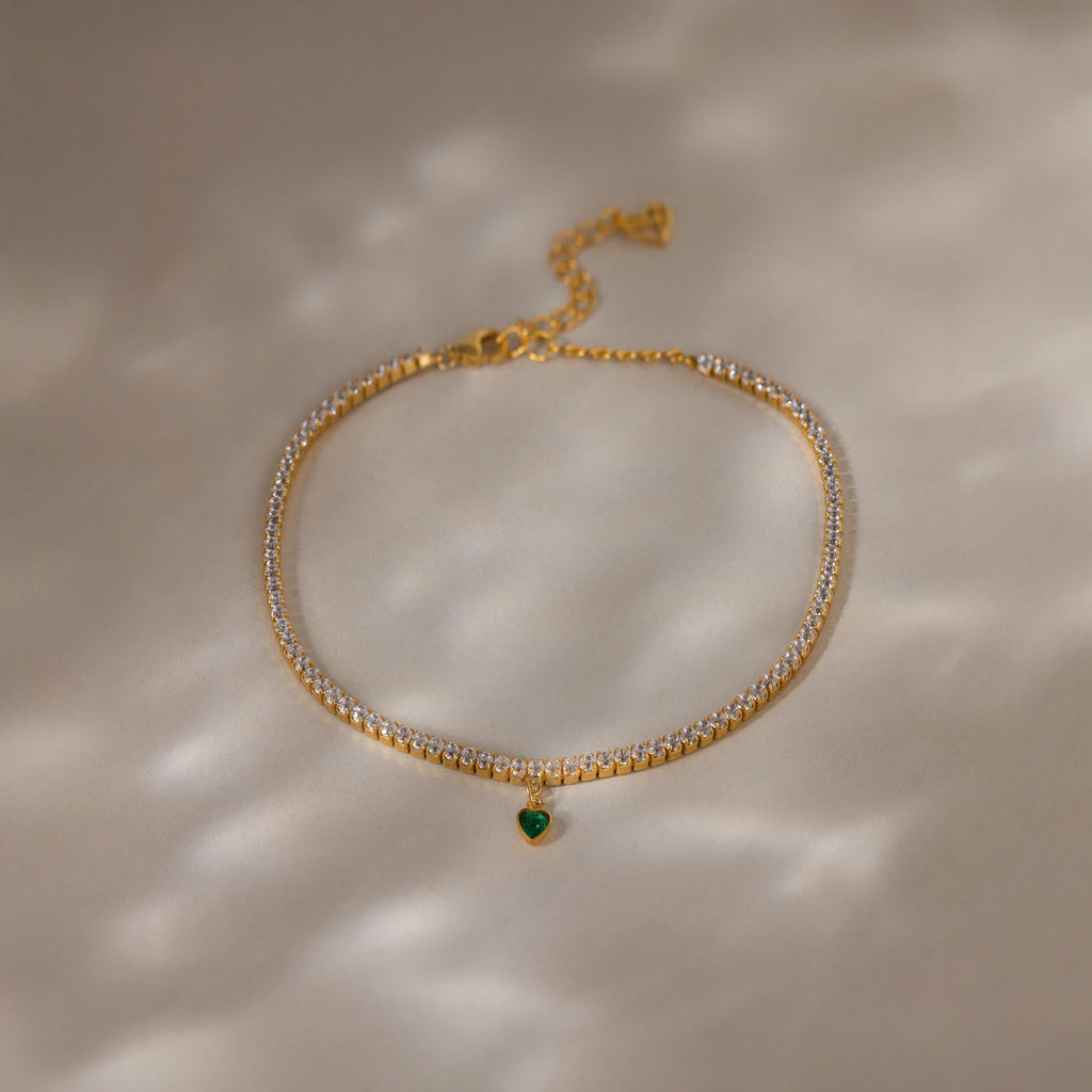 A Heart Birthstone Tennis Bracelet with clear stones and a small green heart charm, reminiscent of personalized or CZ birthstone jewelry, displayed on a beige surface.