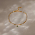 A Heart Birthstone Tennis Bracelet with clear stones and a small green heart charm, reminiscent of personalized or CZ birthstone jewelry, displayed on a beige surface.