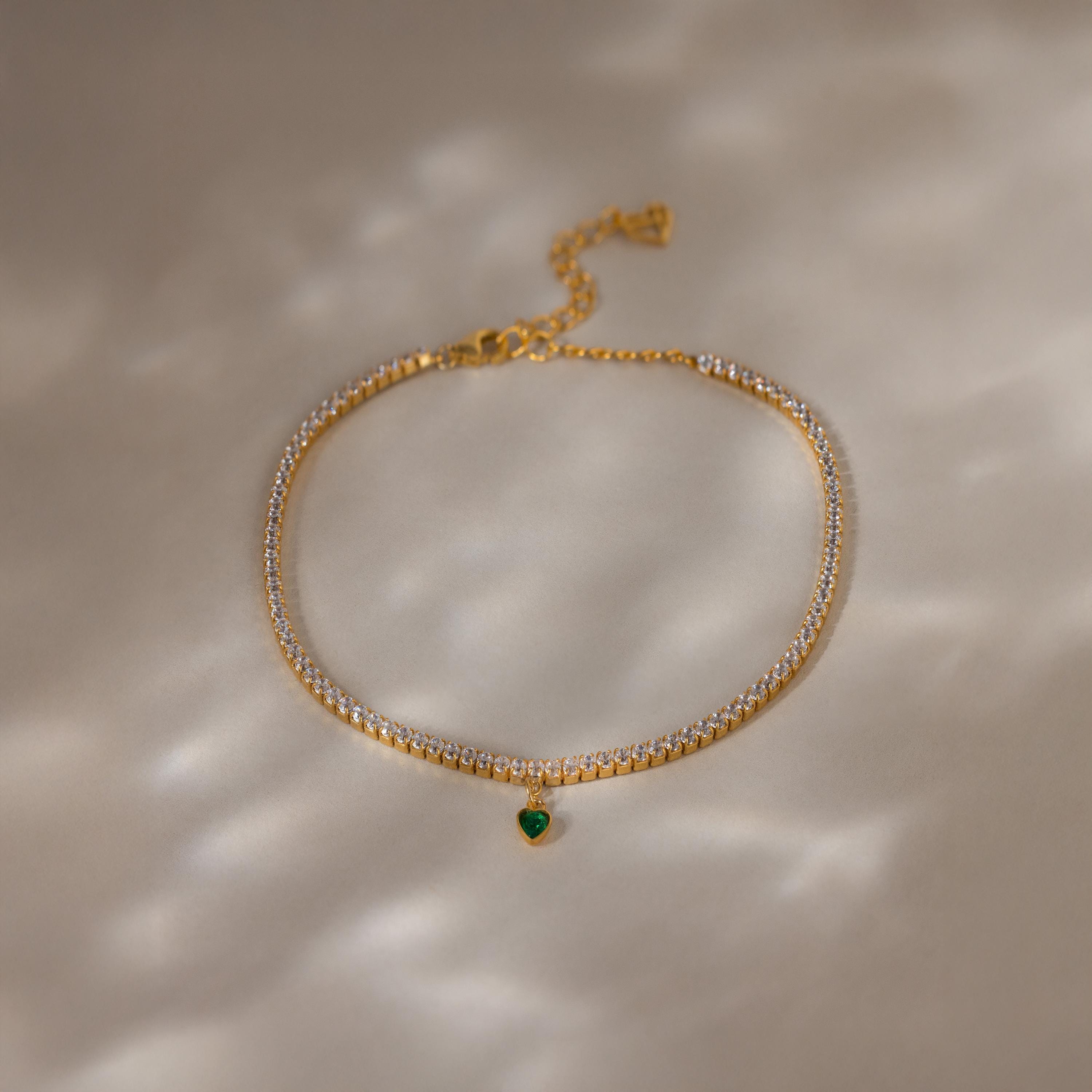 A Heart Birthstone Tennis Bracelet with clear stones and a small green heart charm, reminiscent of personalized or CZ birthstone jewelry, displayed on a beige surface.