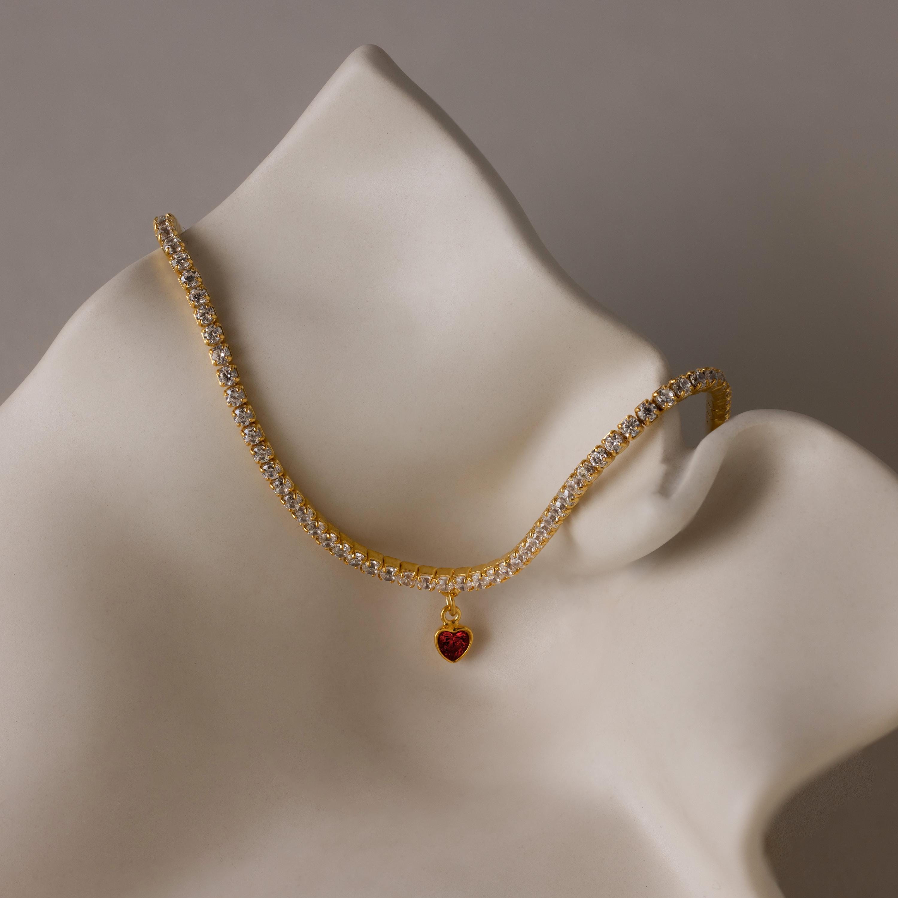 The Heart Birthstone Tennis Bracelet, featuring a gold band with clear stones and a red heart charm, is elegantly displayed on a white abstract sculpture.
