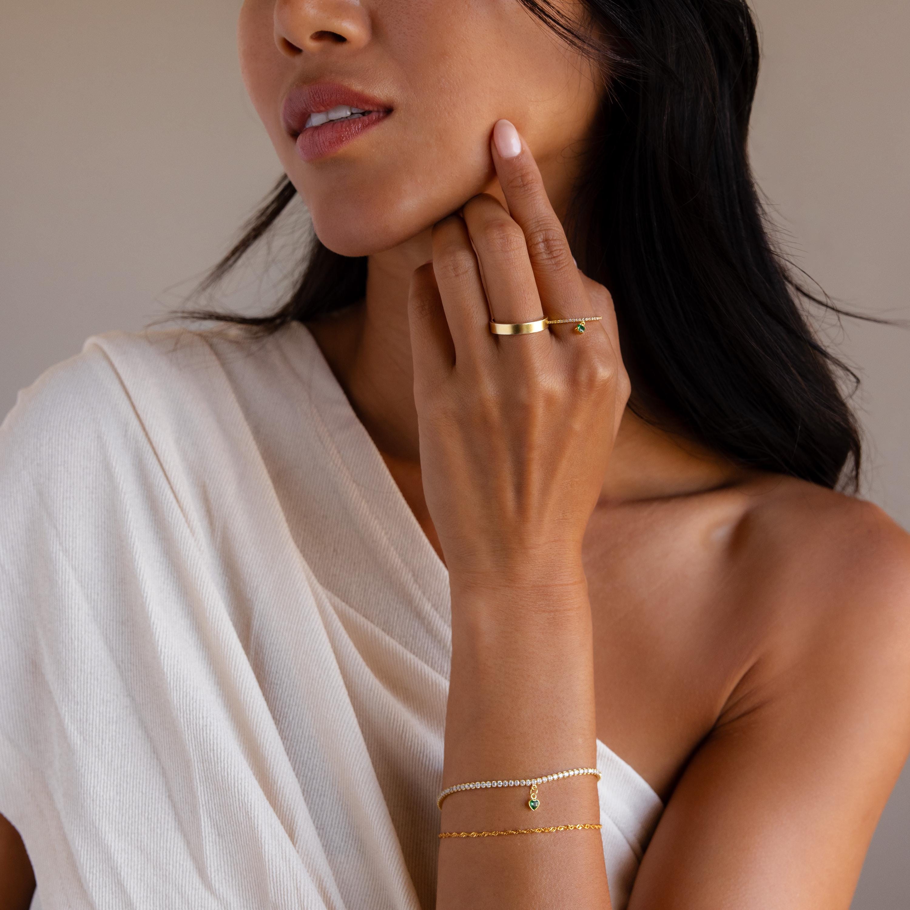 Woman in an off-white top wears gold rings, a gold bracelet, and the Heart Birthstone Tennis Bracelet featuring pearls and a green CZ stone, exuding elegance and charm.