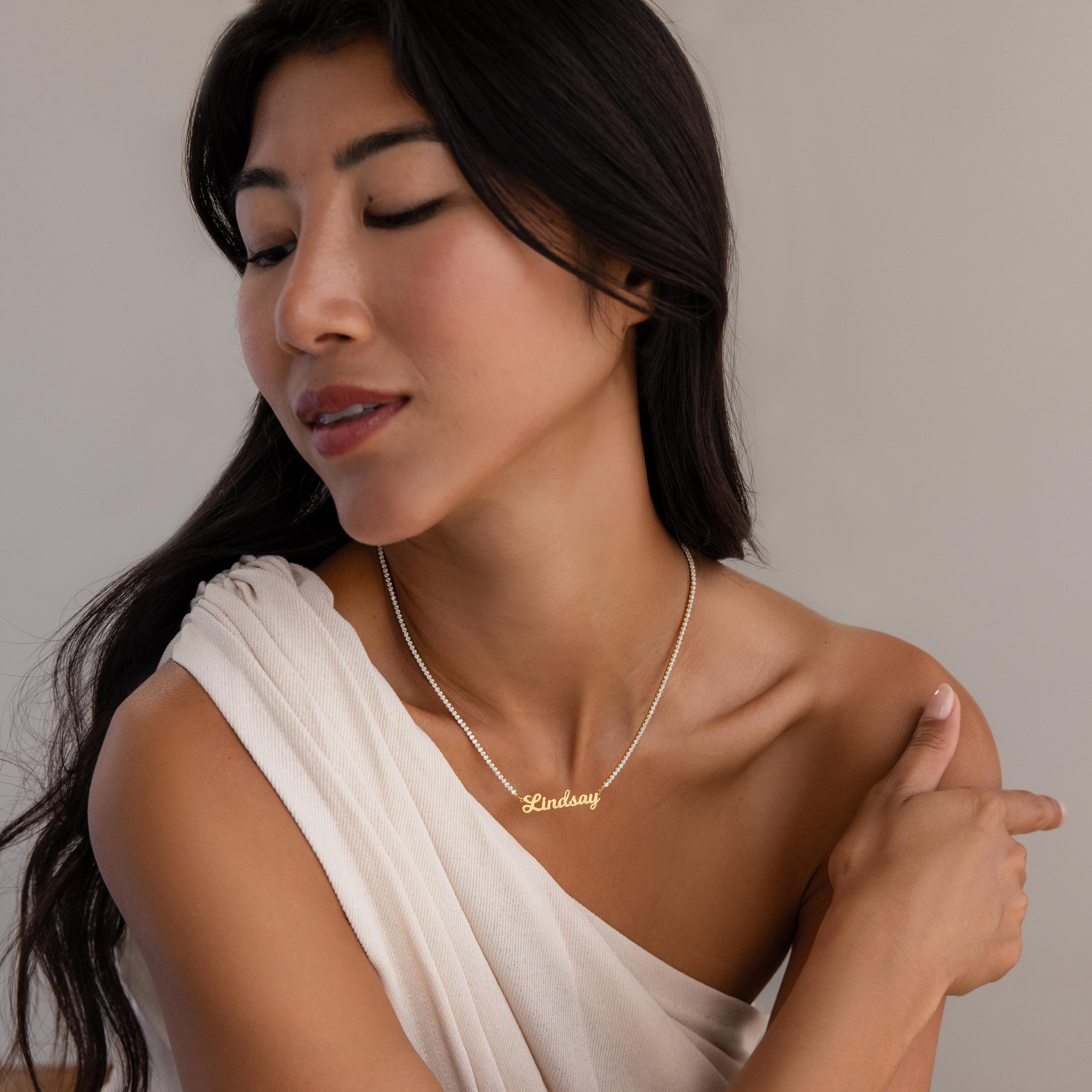 A woman in a white off-shoulder top looks down and gently touches her shoulder while wearing the Diamond Tennis Name Necklace personalized with "Lindsay.