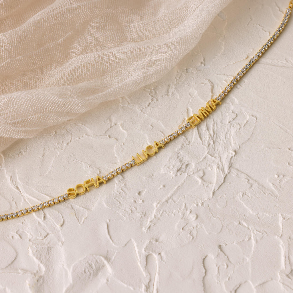 The Multiple Name Tennis Bracelet featuring SOFIA, LUCA, and EMMA rests on a textured white surface with beige fabric—an elegant way to display stylish custom name charms.