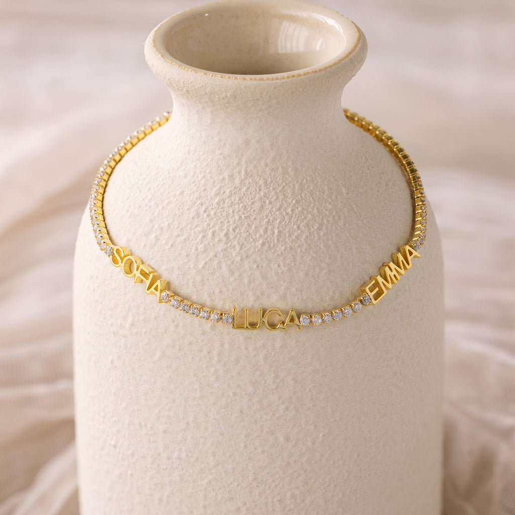 The Multiple Name Tennis Bracelet, shown with the names SOFIA, LUCA, and EMMA on a textured cream vase, is perfect for pairing with custom name charms or as a personalized jewelry statement.