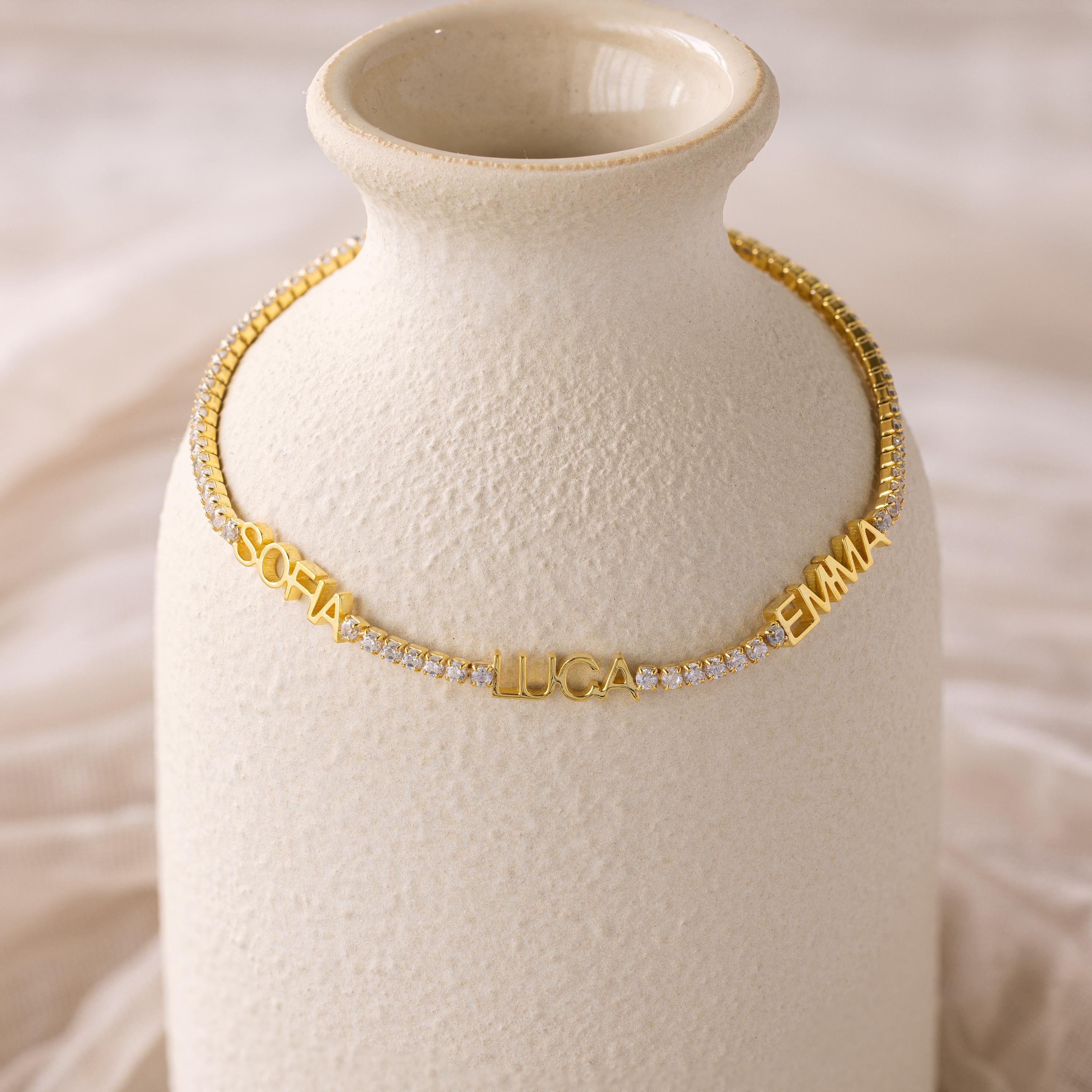 The Multiple Name Tennis Bracelet, shown with the names SOFIA, LUCA, and EMMA on a textured cream vase, is perfect for pairing with custom name charms or as a personalized jewelry statement.