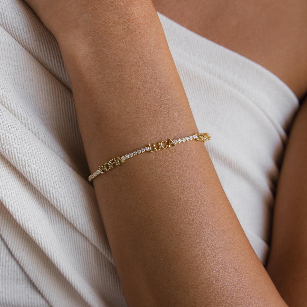 The Multiple Name Tennis Bracelet, featuring custom names, is shown on a person's arm alongside a white textured garment.