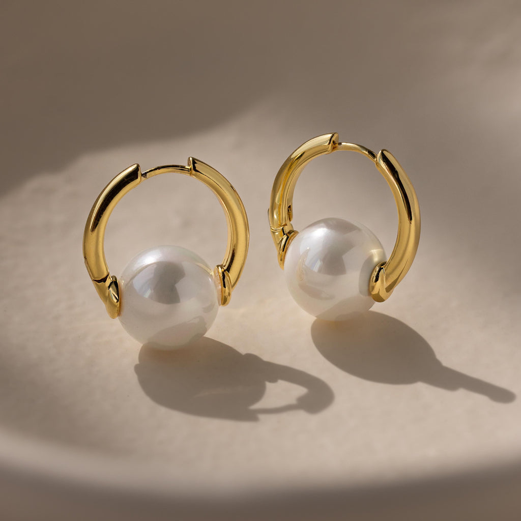 The Bold Round Pearl Hoops showcase gold hoops adorned with large white pearls, displayed on a smooth, light-colored surface under soft lighting.