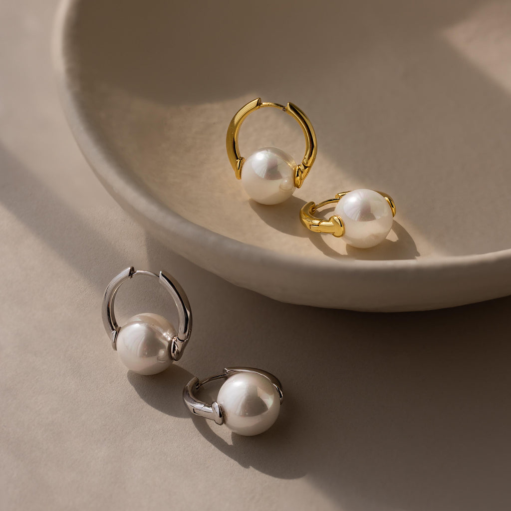 Two pairs of Bold Round Pearl Hoops—one gold, one silver—rest on a neutral ceramic dish, providing timeless style for lovers of statement earrings.